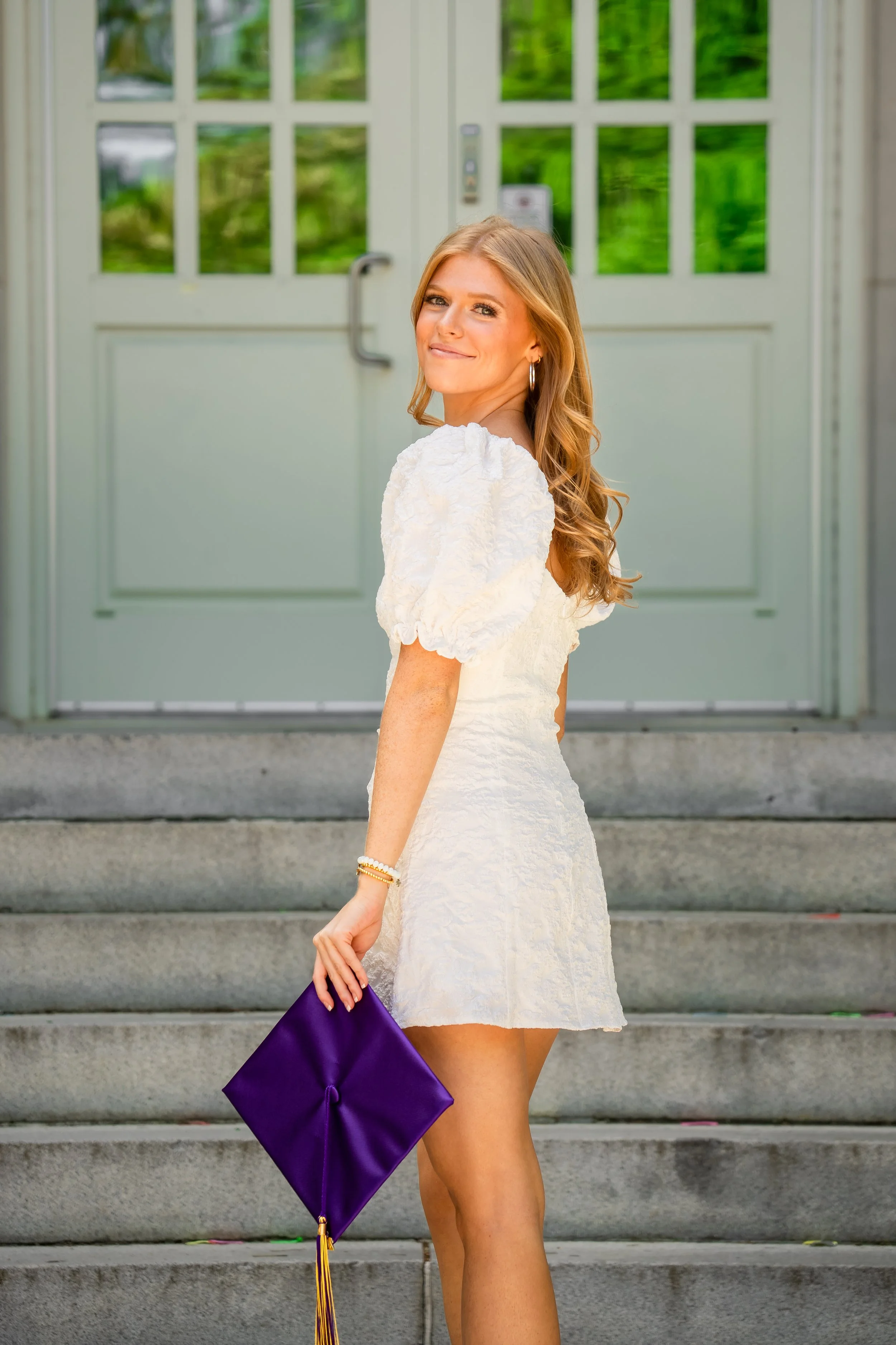 Woman in white graduation dress holding a purple diploma cover, standing on steps in front of a green door.
