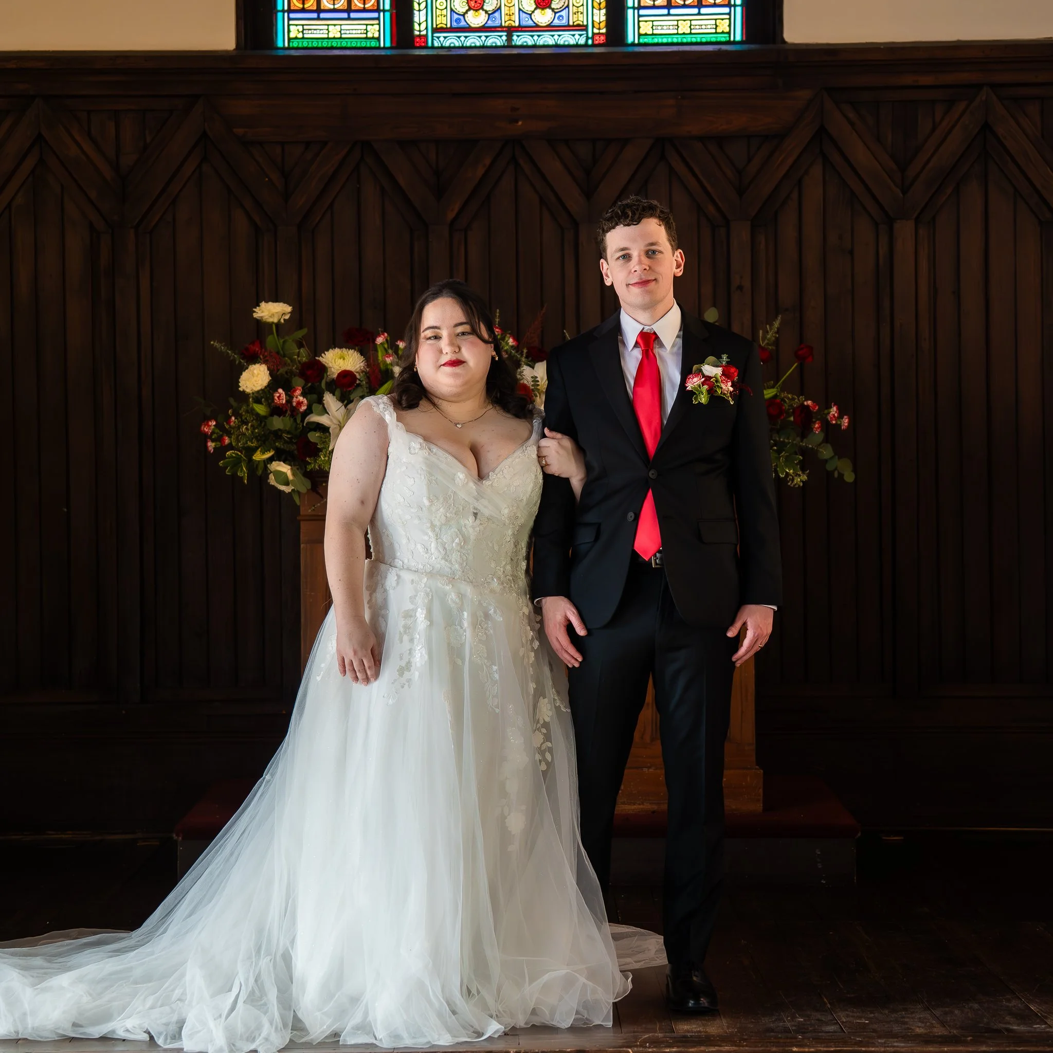 A bride and groom standing together in a church during their wedding ceremony, with a flower arrangement behind them.