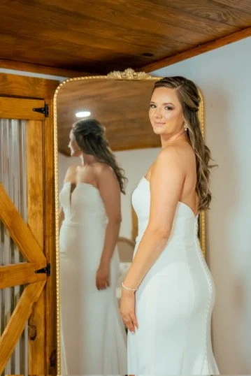 A woman in a white strapless dress stands in front of a mirror, looking at her reflection.