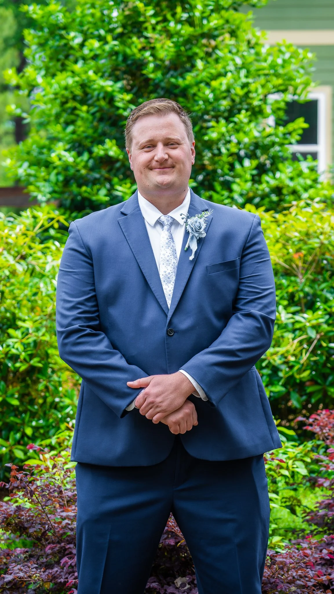 A man in a navy blue suit with a boutonniere stands outdoors in front of a green bush, smiling.