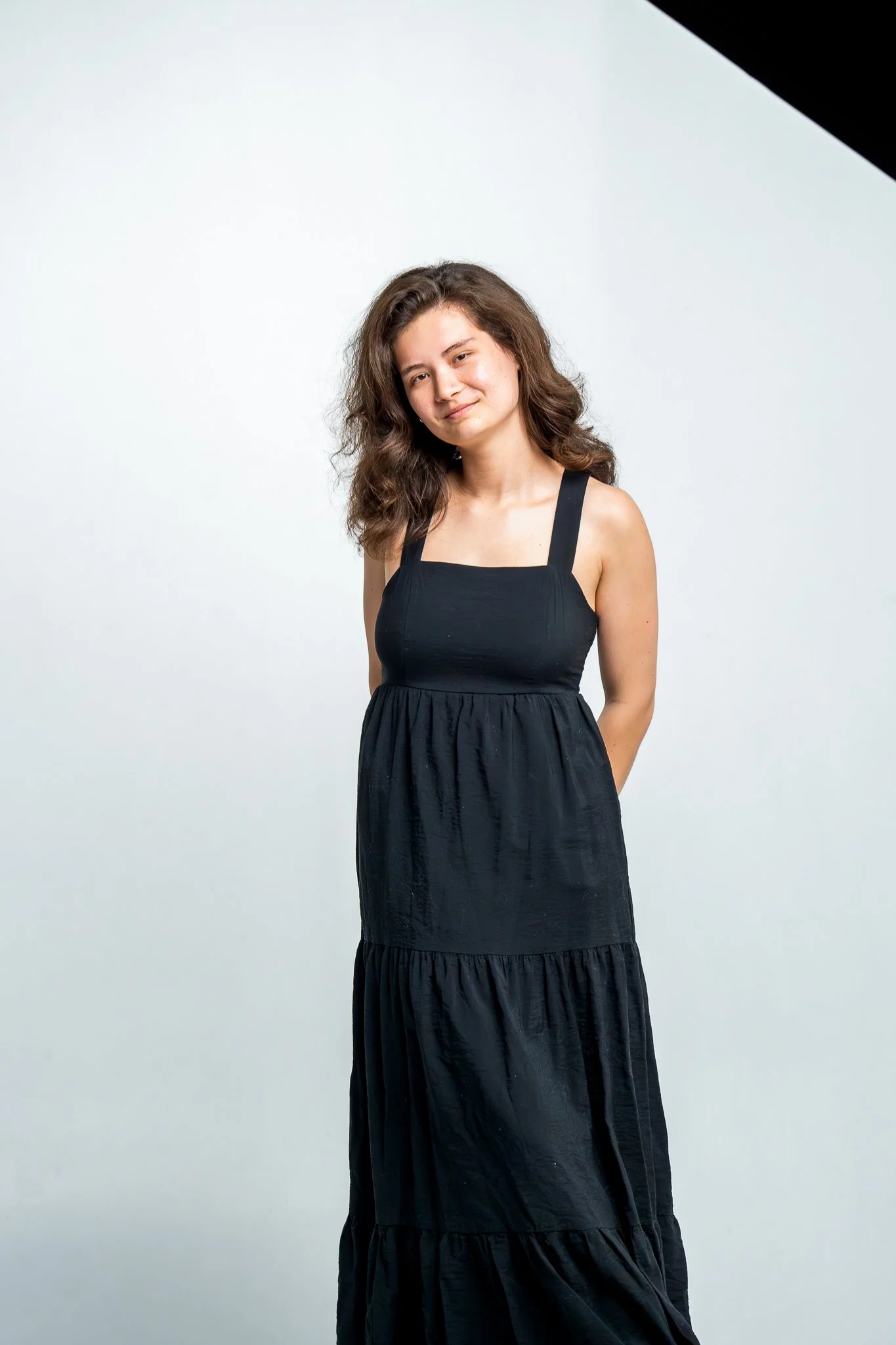 Portrait of a young woman with wavy brown hair wearing a black sleeveless dress, standing against a white background.