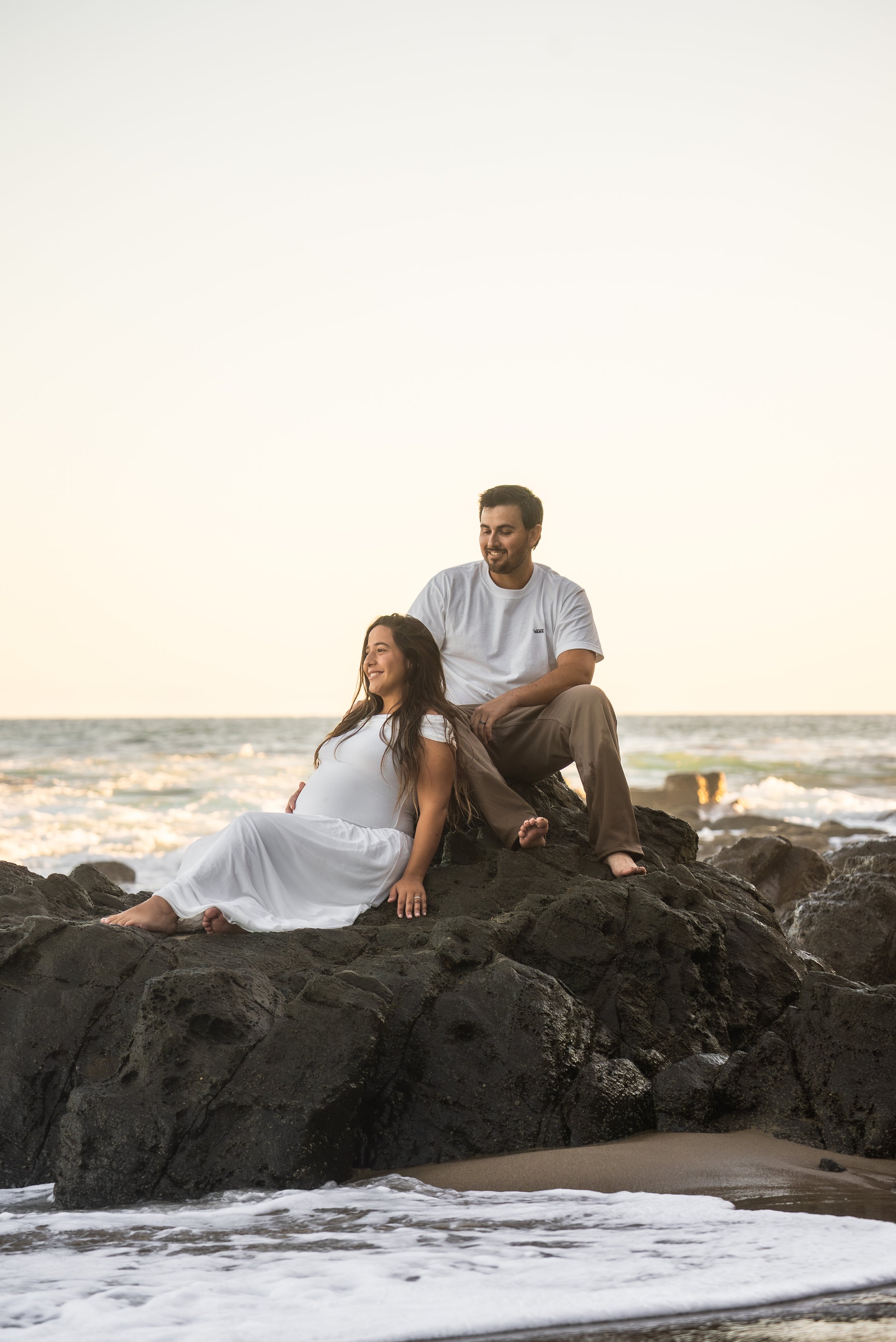 A man and woman sitting on dark rocks at the beach during sunset; the woman is in a white dress and lying back, smiling, the man is in a white T-shirt and khaki pants, sitting behind her, smiling, looking at her.
