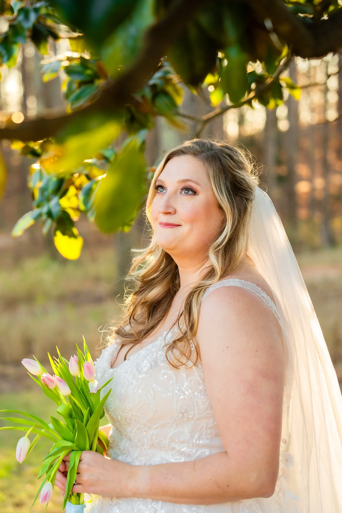 Bride with long blond hair in a white lace wedding dress holding a bouquet of pink tulips, standing outdoors in a forest setting during sunset.
