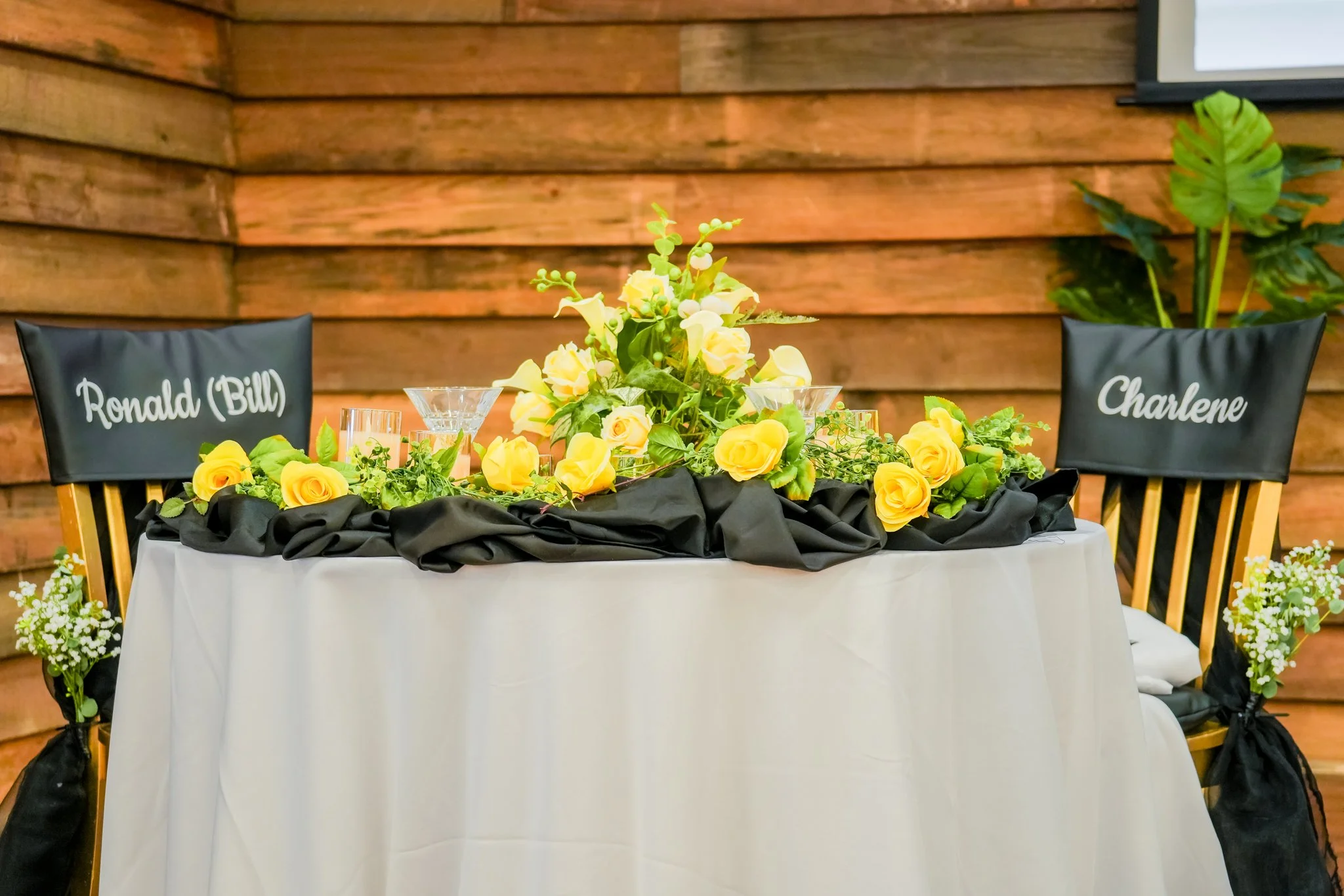 Table with black and white cloth, decorated with yellow flowers, two chairs with name tags 'Ronald (Bill)' and 'Chalene', and a wooden wall background.