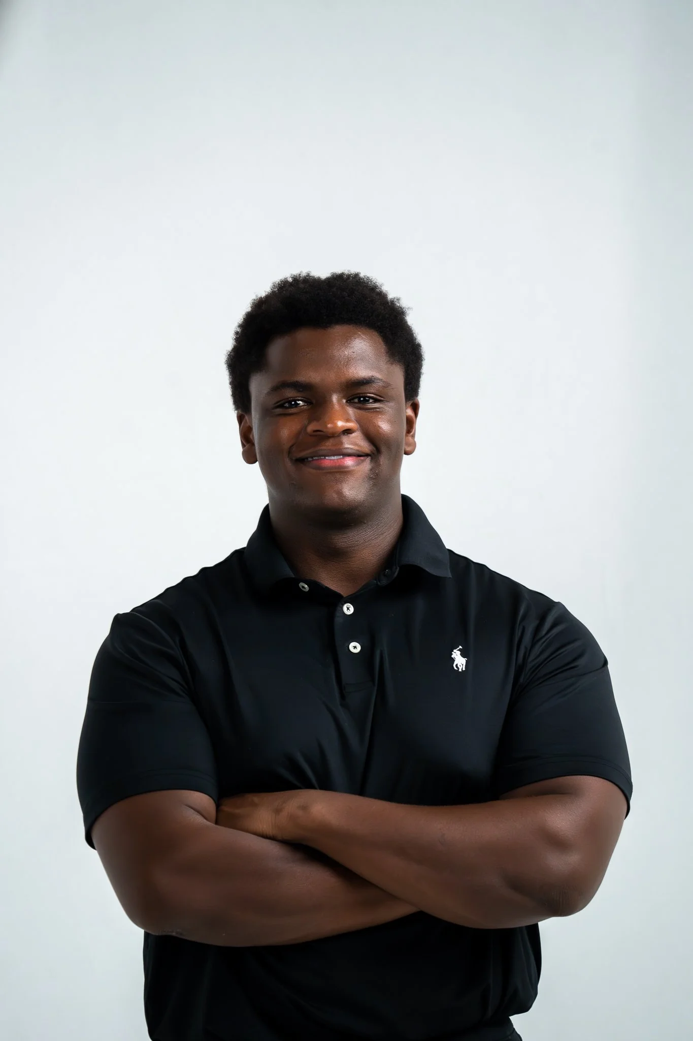A young man with dark skin and curly hair stands against a plain white background, smiling, wearing a black polo shirt with a small white logo on the left chest, arms crossed.