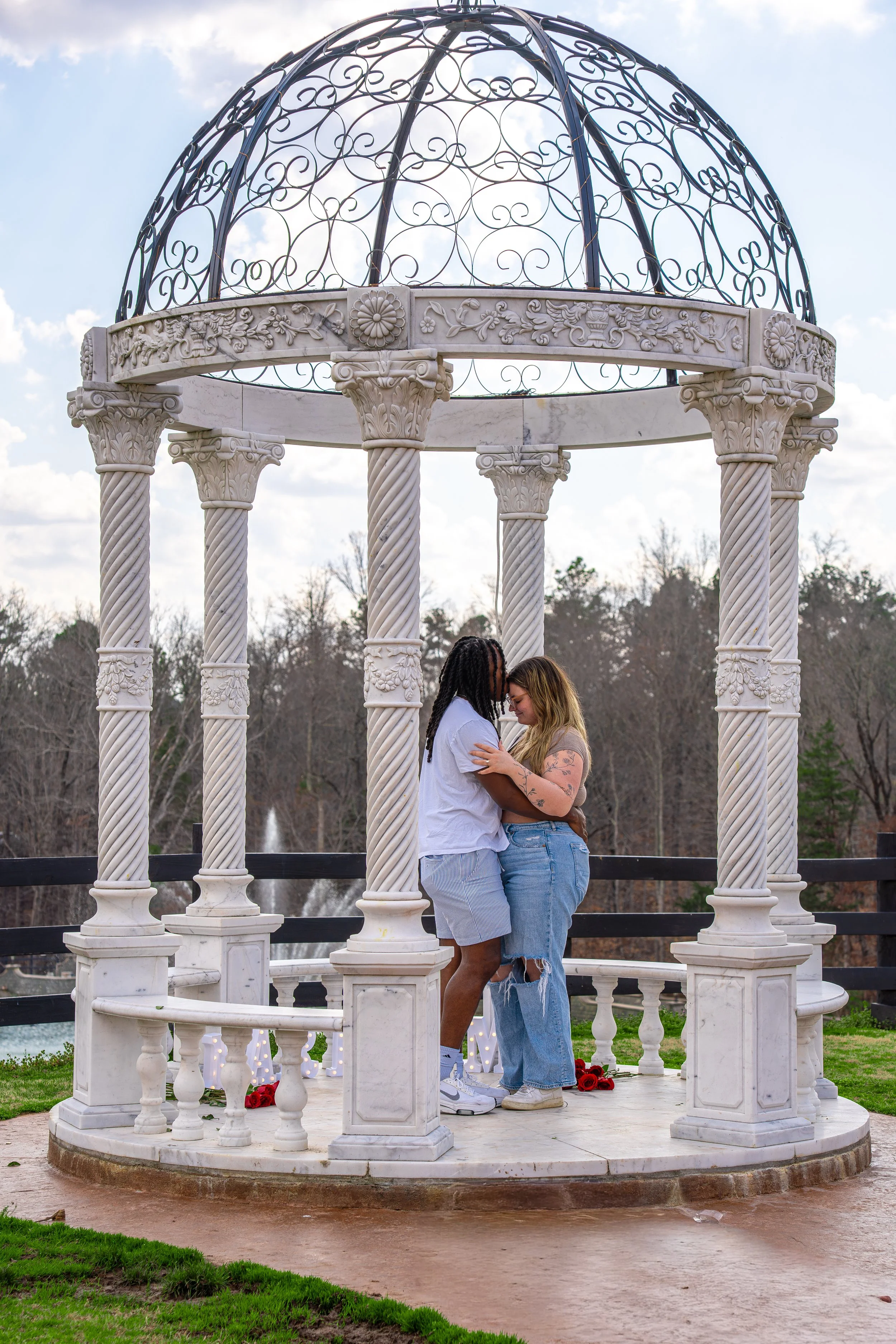 Two people are standing close together inside a white marble gazebo with ornate columns and a decorative iron dome top. They are touching foreheads affectionately, with a small bouquet of red roses on the ground nearby, and trees and a fountain in the background.