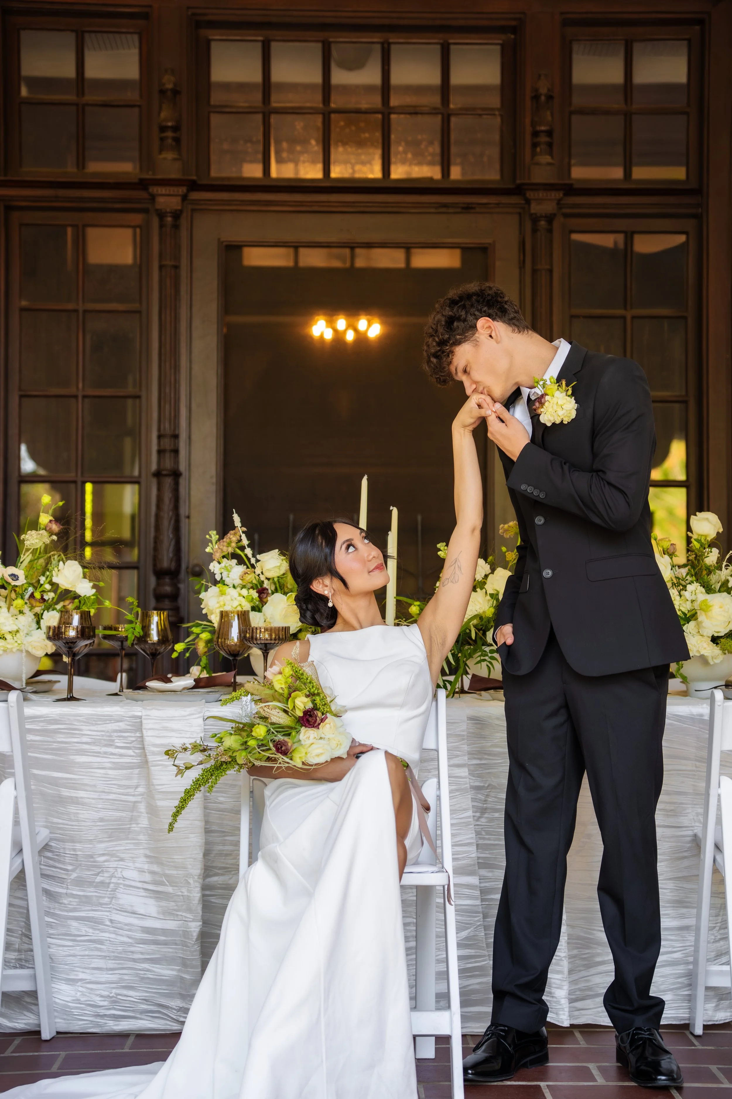 Bride seated at the reception table while the groom kisses her hand during an elegant wedding reception celebration.
