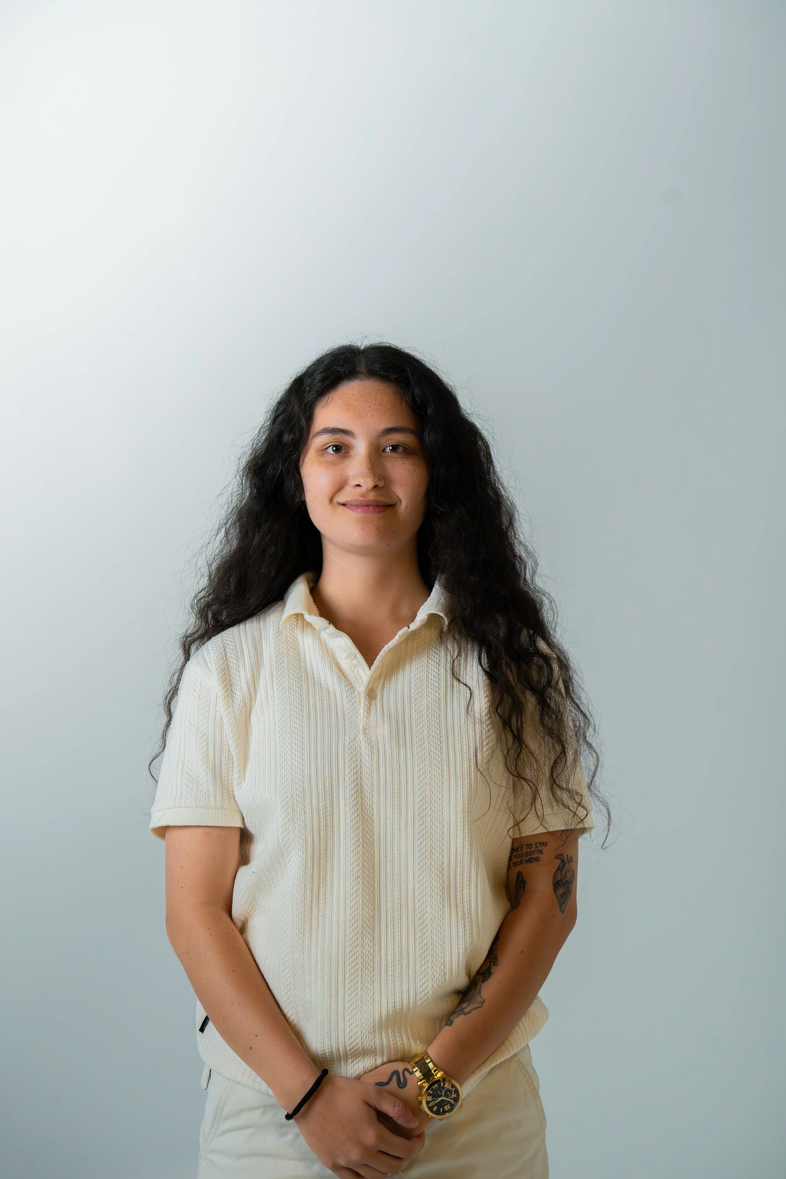 Young woman with long curly dark hair wearing a cream polo shirt and beige pants, standing against a plain light gray background.