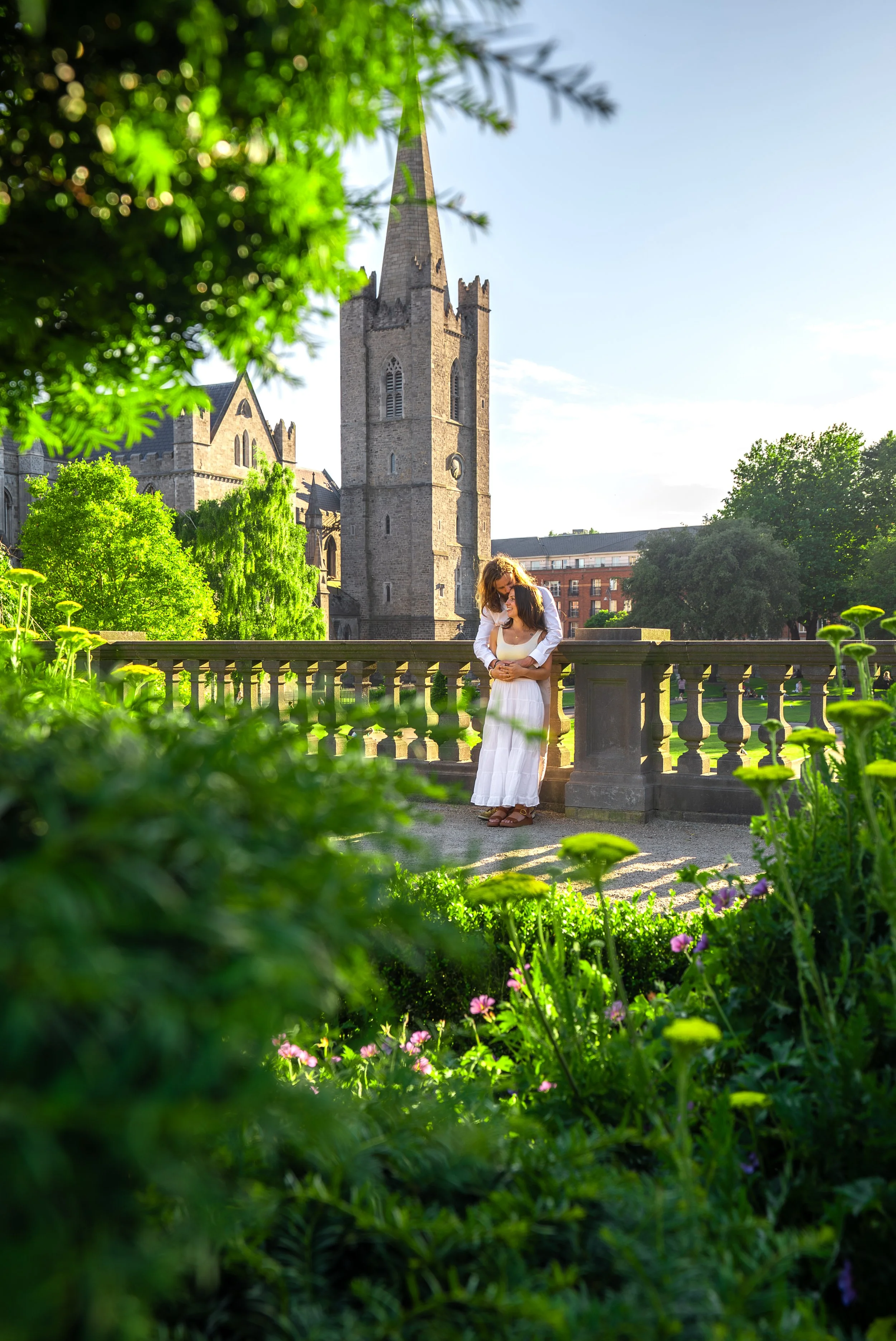 Two women hugging on a stone balcony in a garden with green trees and a church in the background. The sky is clear and the lighting suggests late afternoon or early evening.