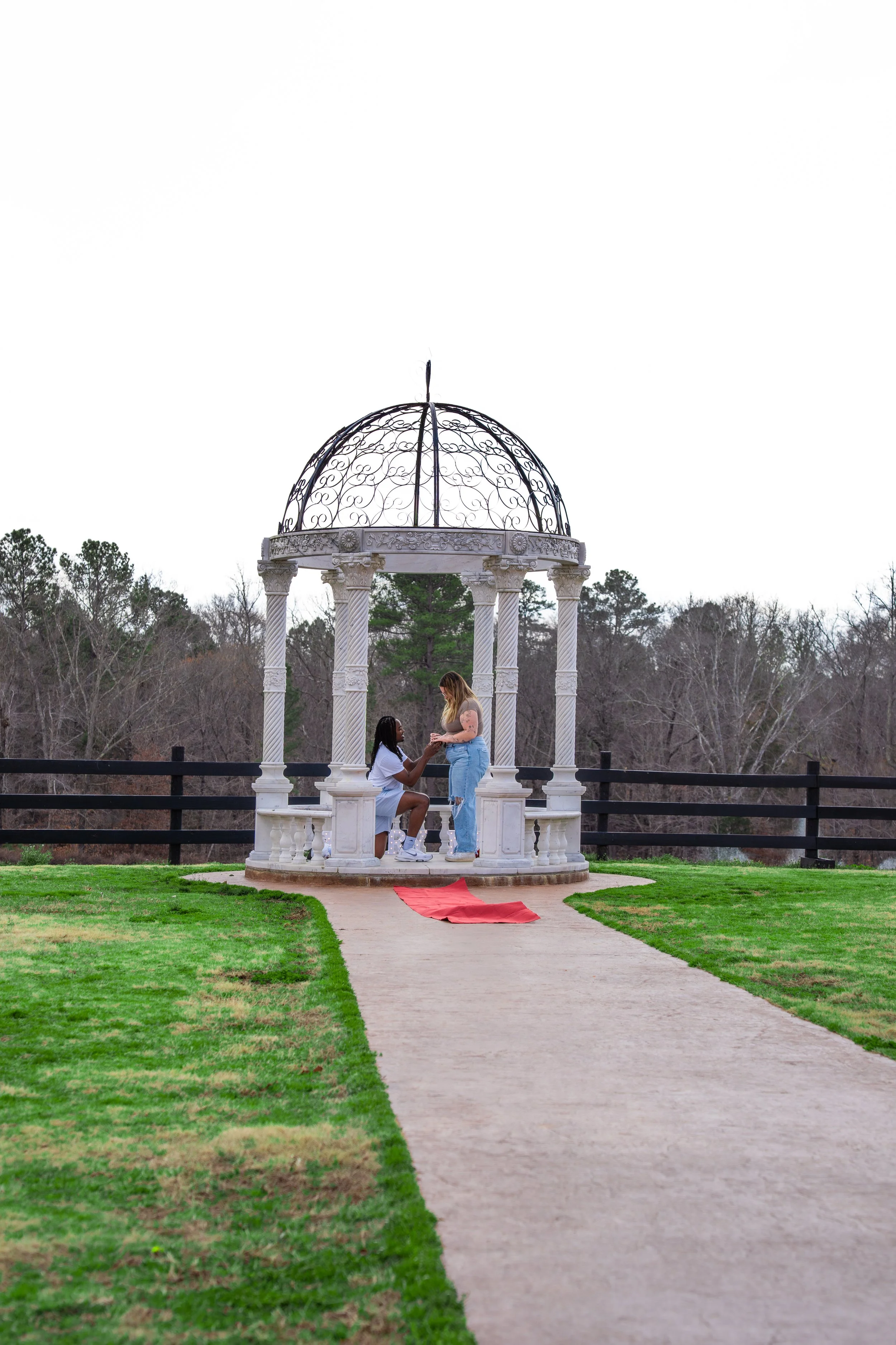 Two women exchanging a ring inside a white, ornate gazebo on a grassy area, with trees and a black fence in the background.