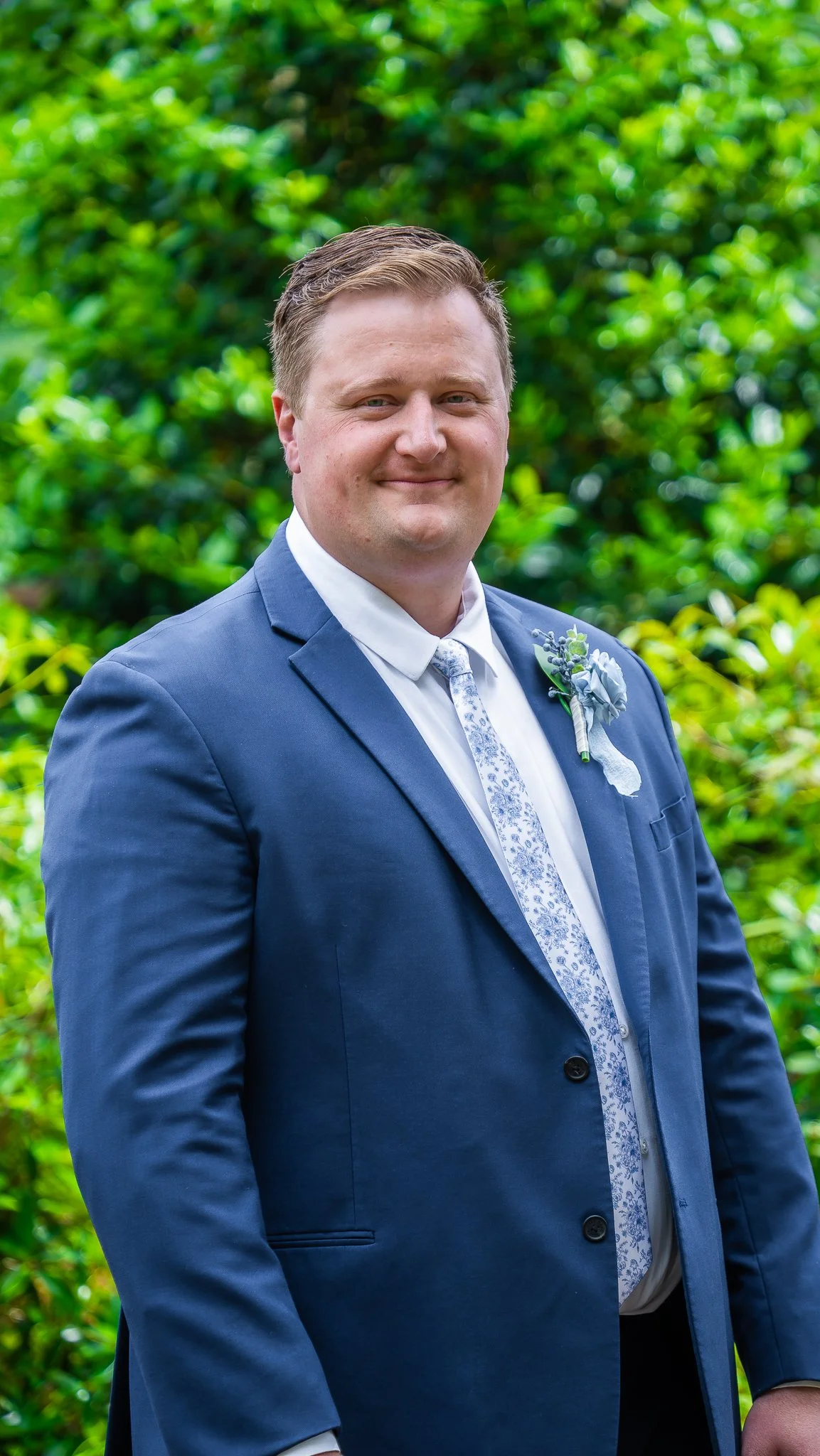 A man in a blue suit standing outdoors with greenery in the background, smiling at the camera.