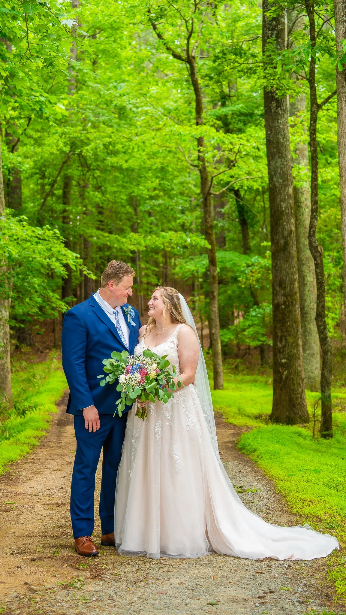 A bride and groom standing closely on a woodland path, smiling at each other. The bride is holding a bouquet of flowers, wearing a white wedding gown and veil. The groom is dressed in a blue suit, white shirt, and tie. Surrounding them are green tree