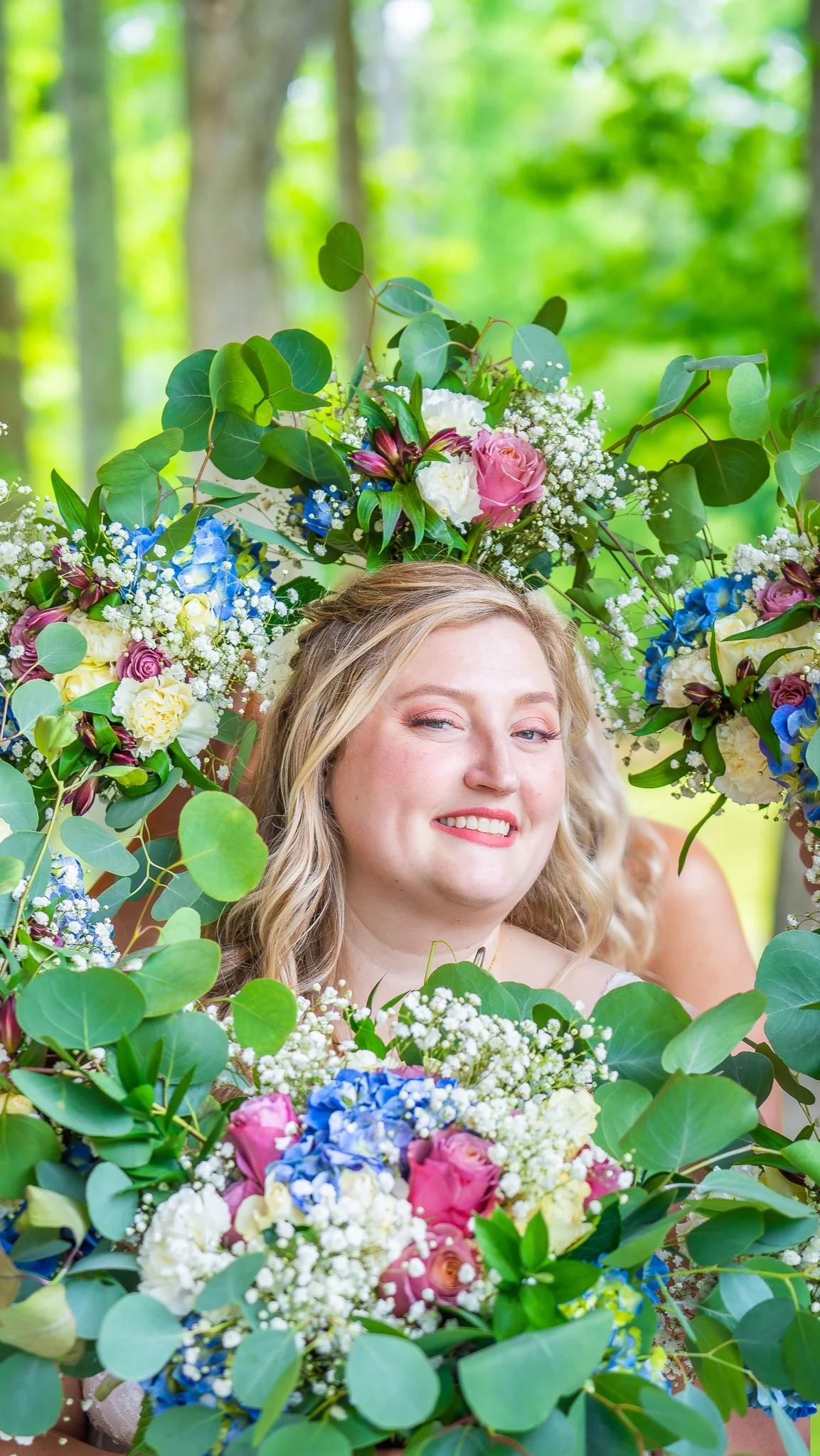 A woman with blonde hair and a pink lipstick smirk stands amidst a lush, green outdoor setting, surrounded by colorful flowers, leaves, and a floral arrangement creating a partial wreath around her head and shoulders.