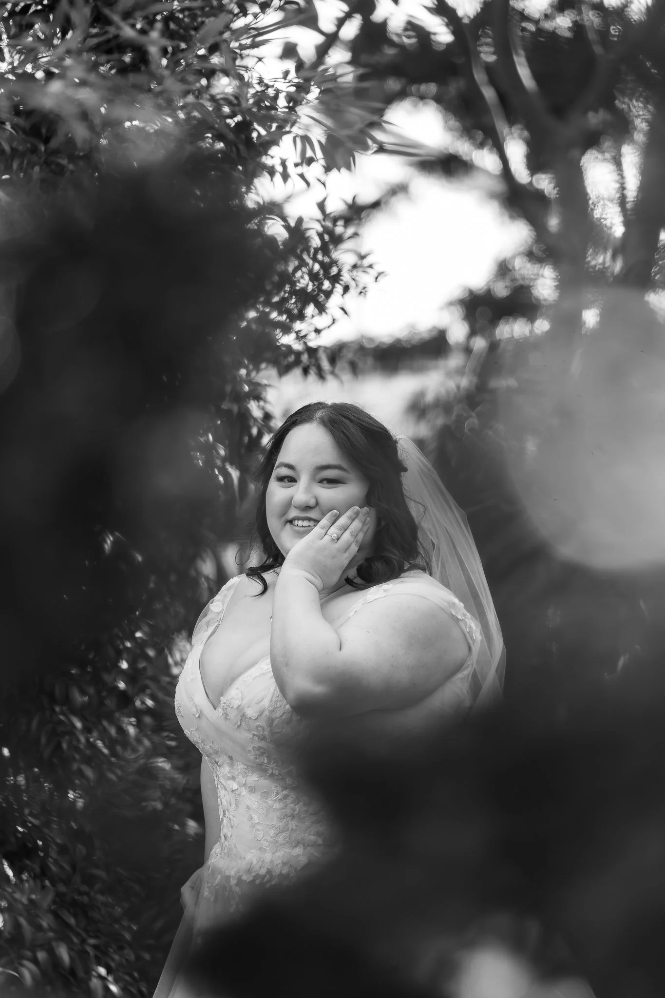 Black and white portrait of a smiling woman in a wedding dress, with her hand on her cheek showing an engagement ring, surrounded by blurred foliage.