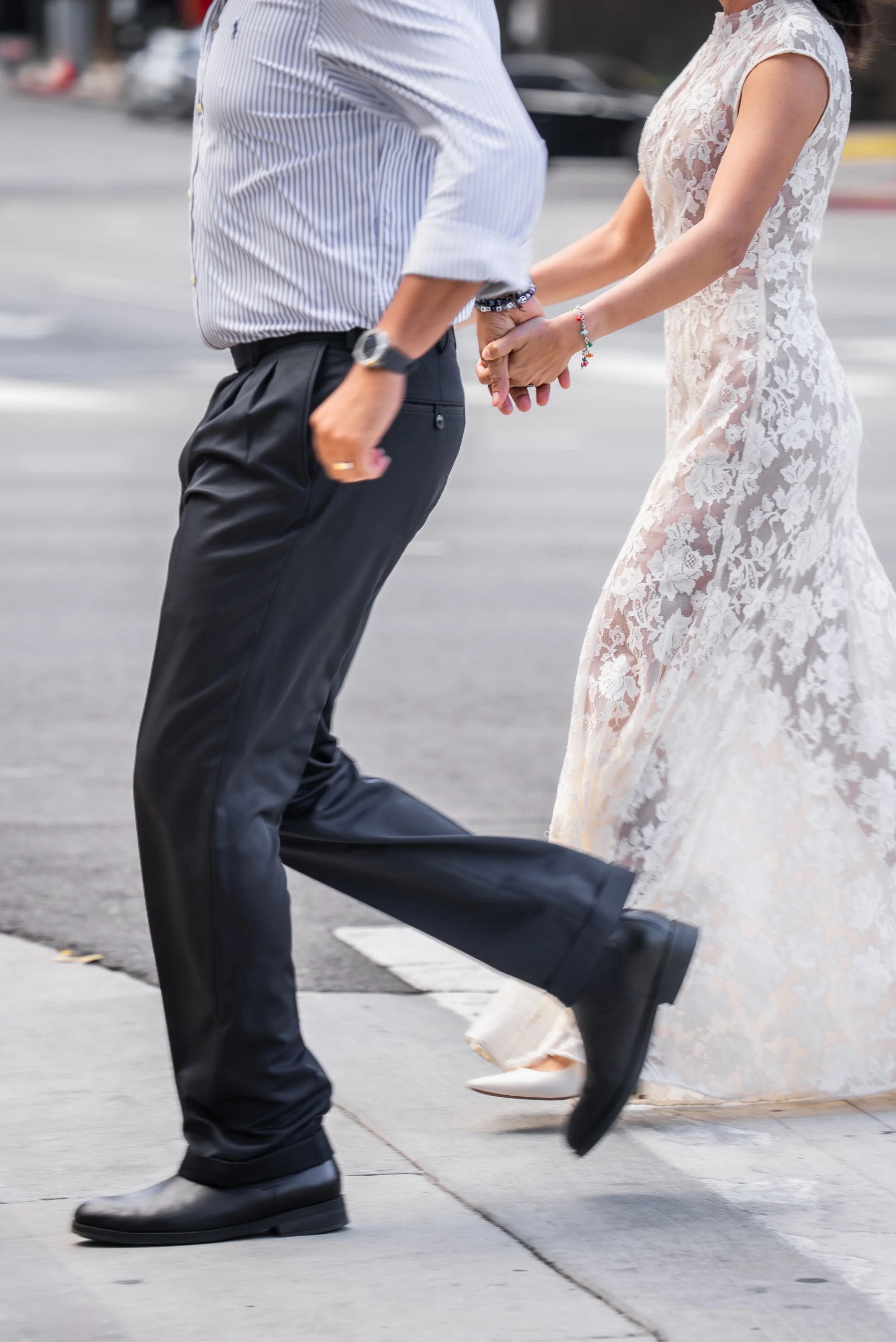 A couple holding hands while walking across a street, with the woman wearing a lace wedding dress and white heels, and the man in dark dress pants and black shoes.