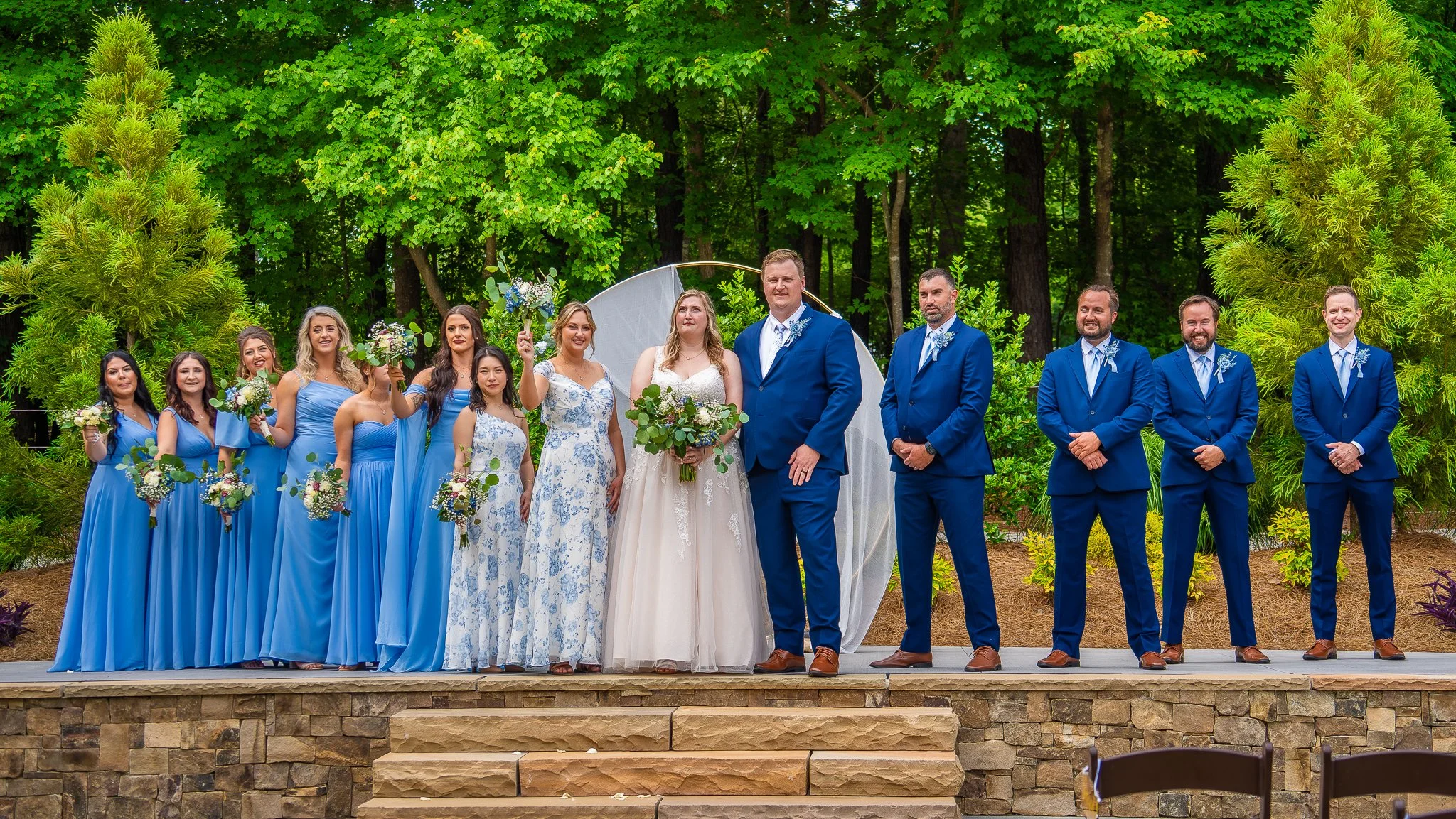 A wedding party standing outdoors on a stone platform, with green trees in the background. The bride and groom are in the center, with the bride holding a bouquet. Bridesmaids dressed in blue and groomsmen dressed in blue suits are positioned on eith