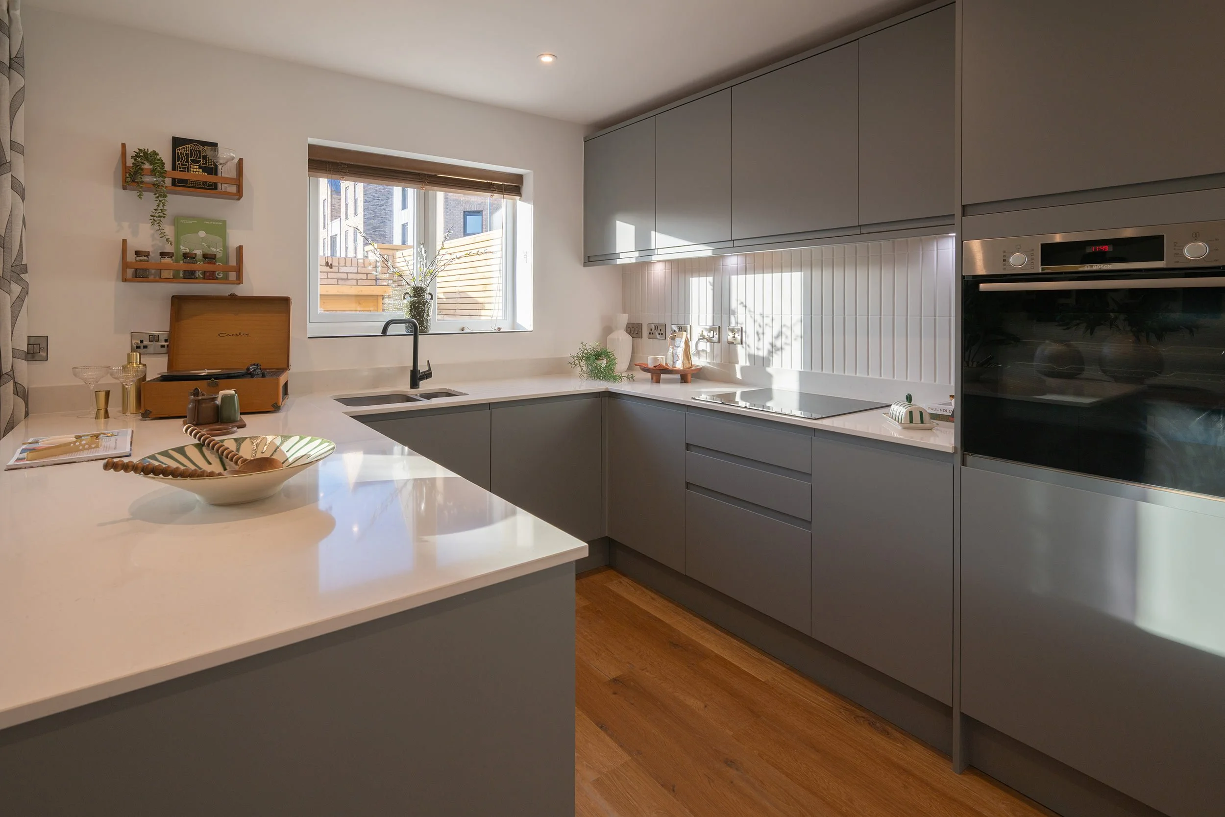 Modern kitchen with gray cabinetry, white countertops, and wooden flooring. Features a window with sunlight, a black faucet, decorative items, and built-in appliances.