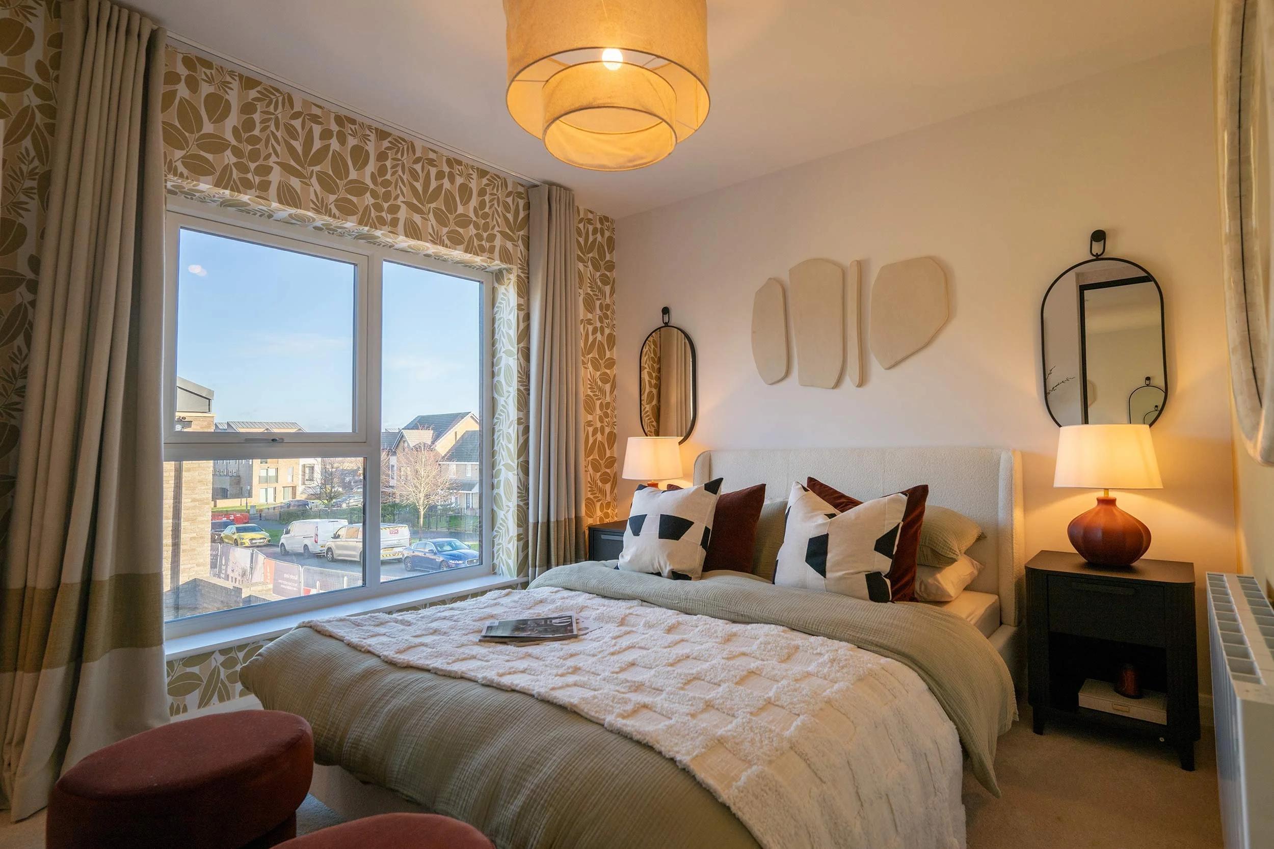 A cozy bedroom with a large window, beige curtains, and patterned wallpaper. The bed has decorative pillows and a textured throw, flanked by two bedside tables with lamps. Wall art and mirrors decorate the wall above the bed.