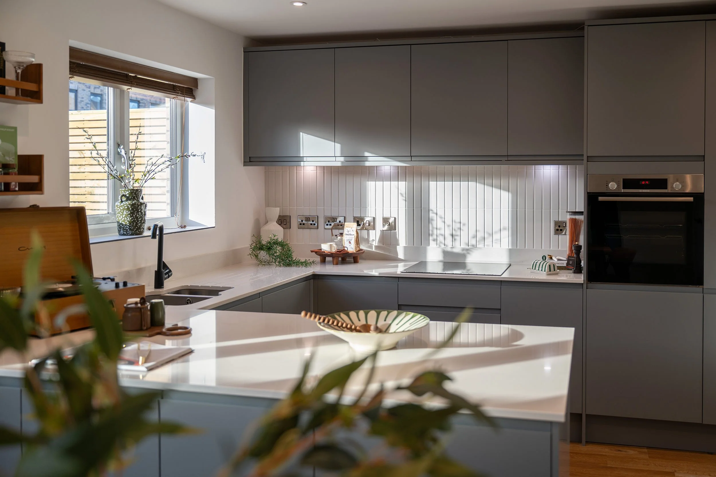 Modern kitchen with gray cabinets, white countertop, and a white backsplash. A window with blinds lets in natural light, and there are decorative items, including a vase with branches, a small plant, and bowls on the counter. An oven is built into th