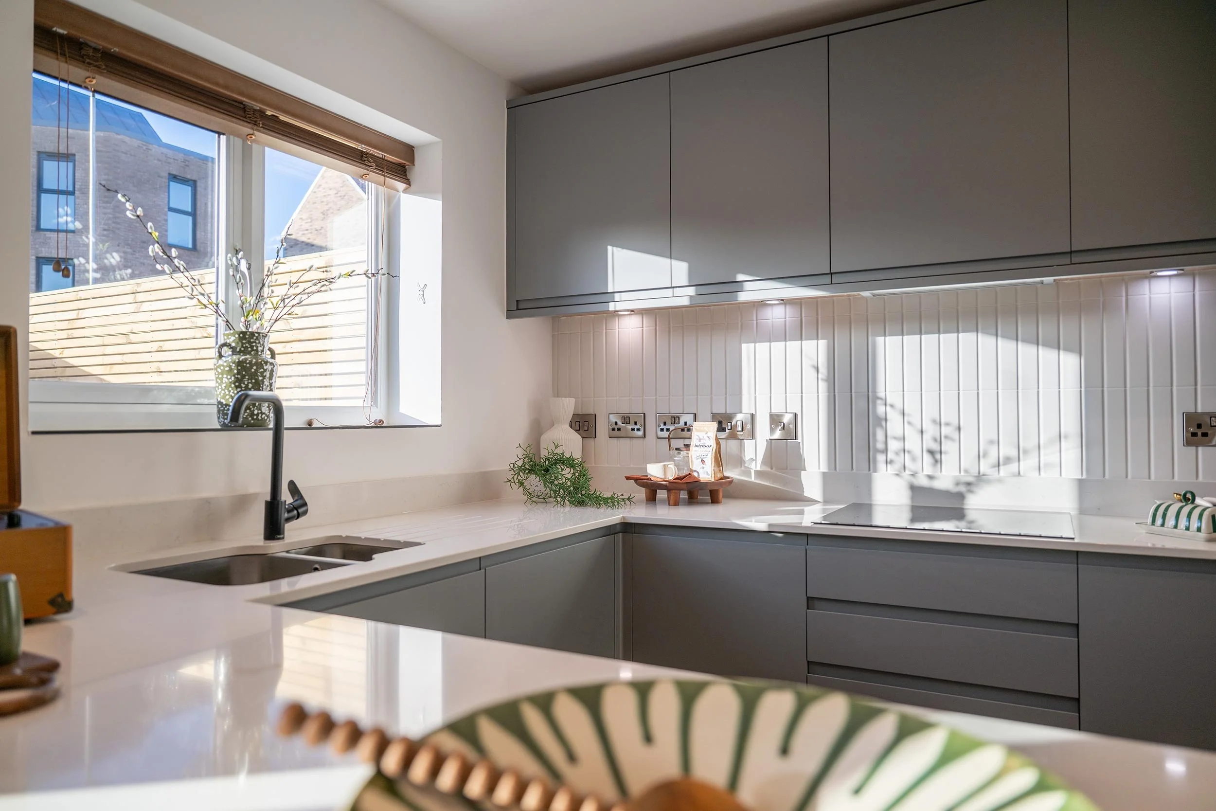 Modern kitchen with white countertops, gray cabinets, a window with a view of a neighboring building, a vase with flowers on the windowsill, and sunlight casting shadows on the backsplash.