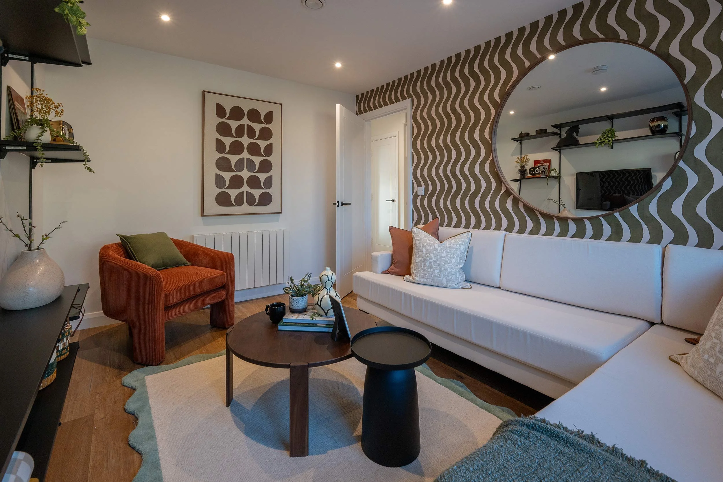Living room with white couch, brown wood coffee table, red armchair with green pillow, modern wall art, and decorative wall mirror with wavy brown and beige pattern.