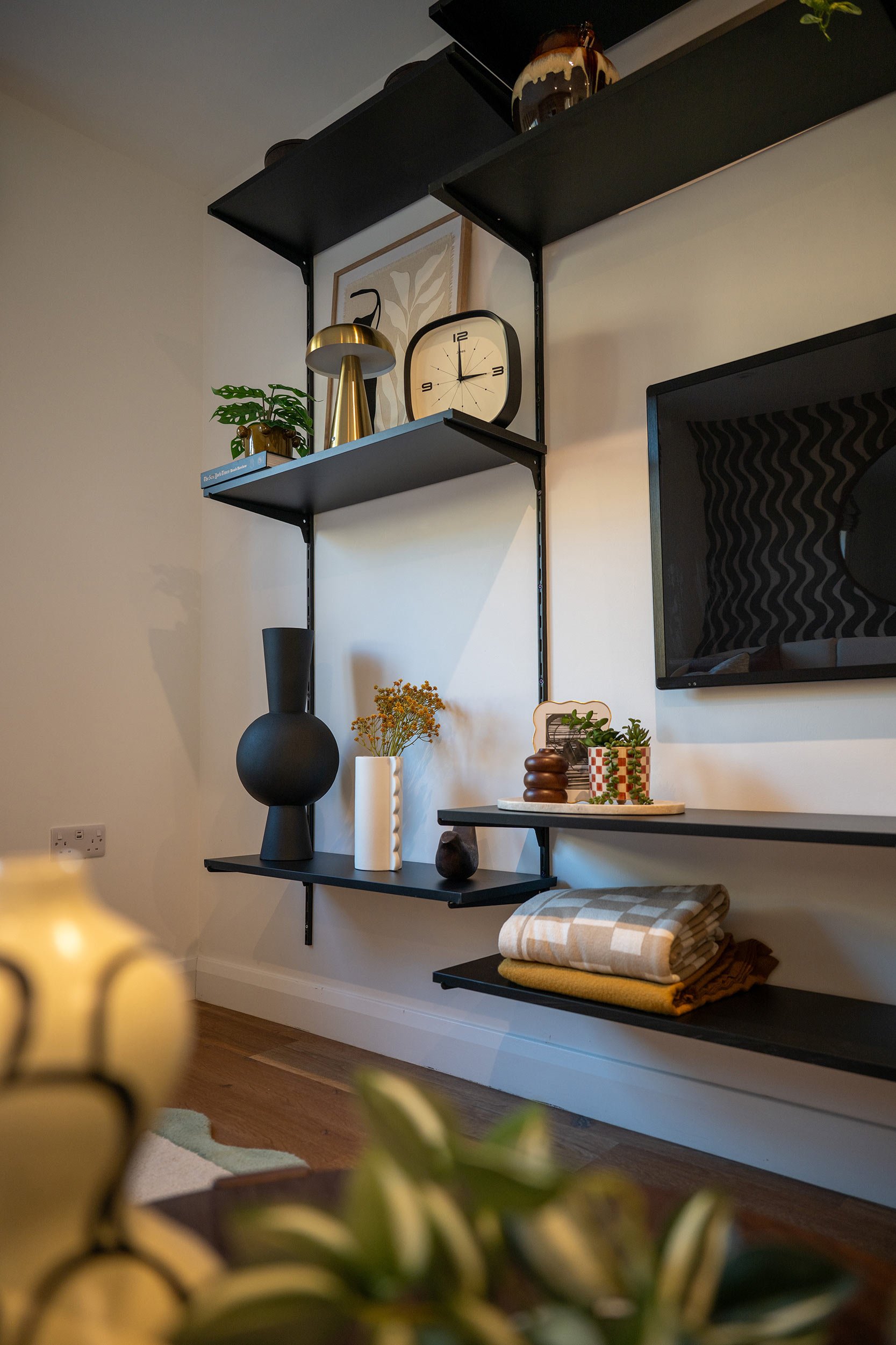 Living room wall with black wall-mounted shelves displaying various decorative items including a clock, vases, plants, and framed artwork.