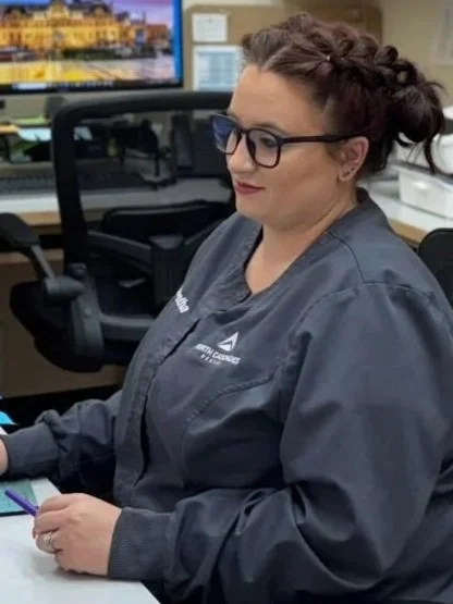 A woman with glasses and braided hair in a gray uniform sitting at a desk with a computer and phone in an office.