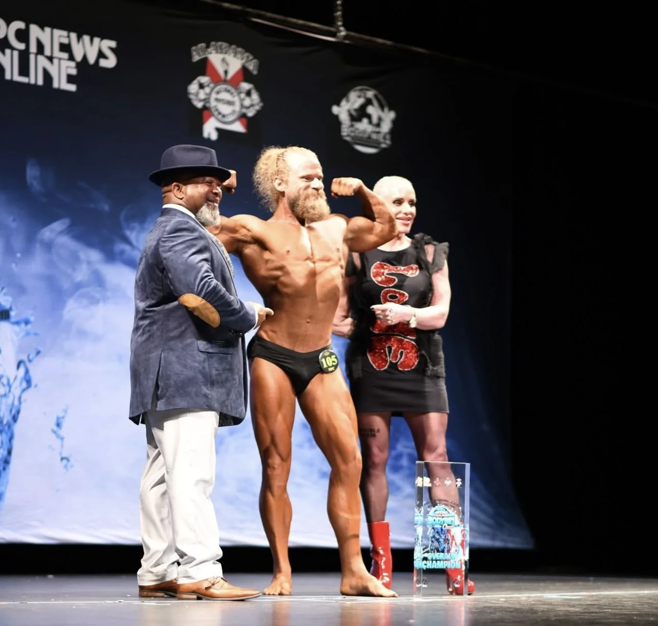 Three people on a stage at a bodybuilding competition, with one man flexing his muscles and the other two smiling, a trophy on the ground, and a dark background with logos and text.