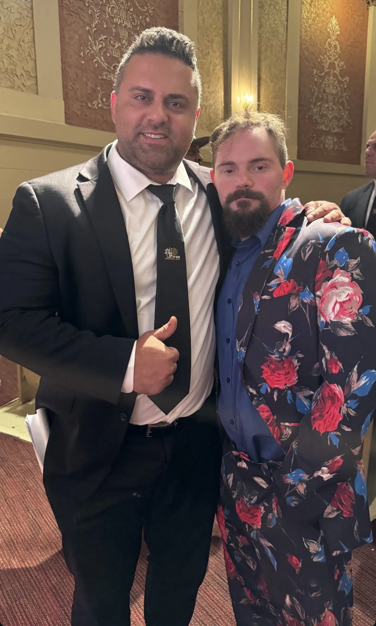 Two men dressed in formal attire posing for a photo indoors, with ornate wall decor and warm lighting in the background.