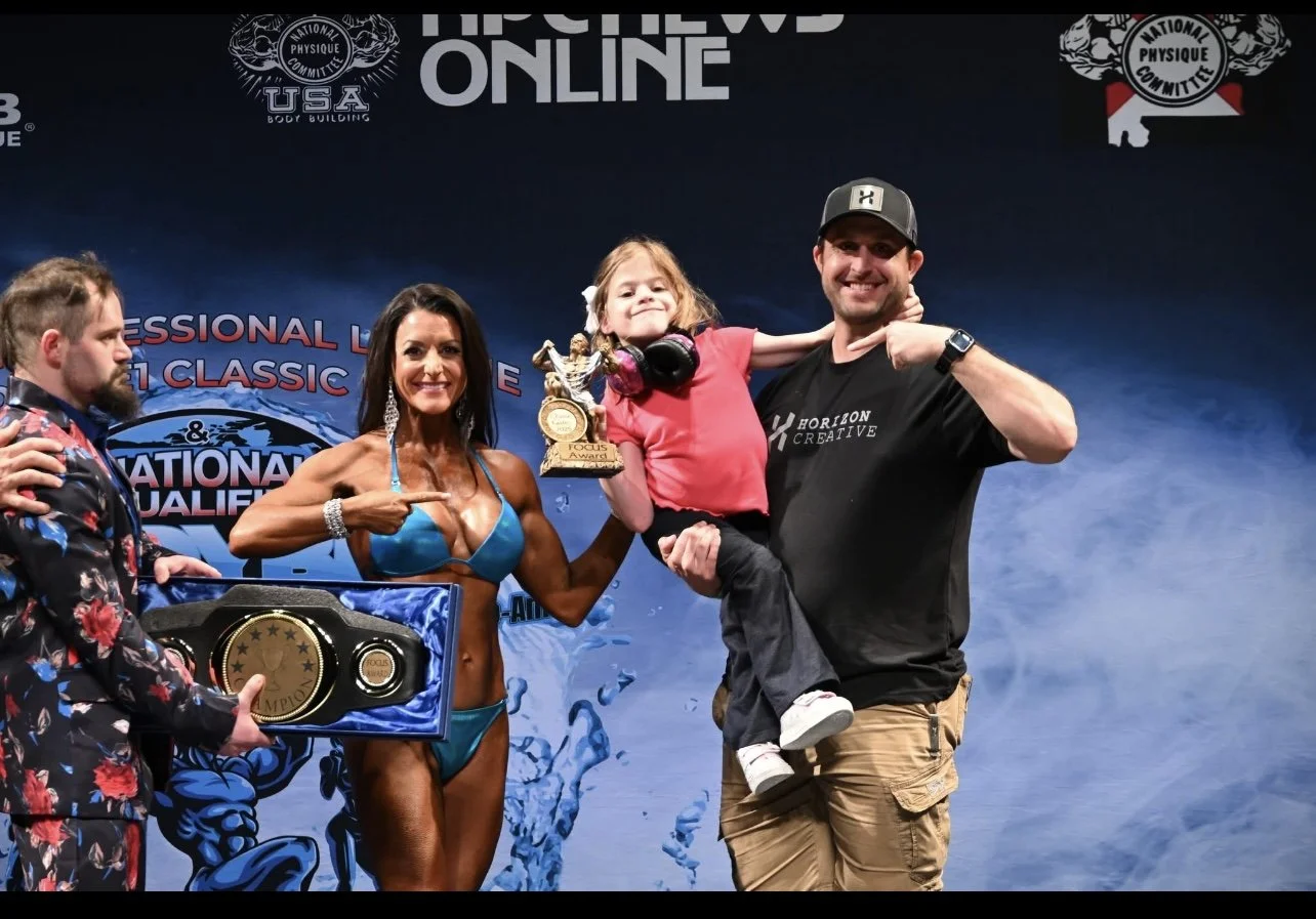 A group of five people on stage celebrating a competition win. A muscular woman in a blue bikini points to a young girl holding a trophy, who is sitting on a man's arm. The man is wearing a black shirt and cap, smiling and flexing his arm. The stage 