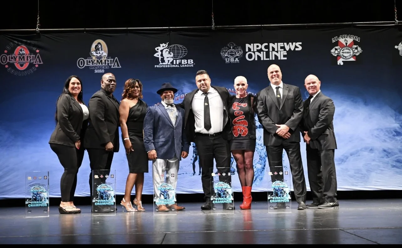 Group of eight people standing on a stage in front of a backdrop with logos, smiling for a photo. Some are holding awards, and one woman is wearing a dress with 