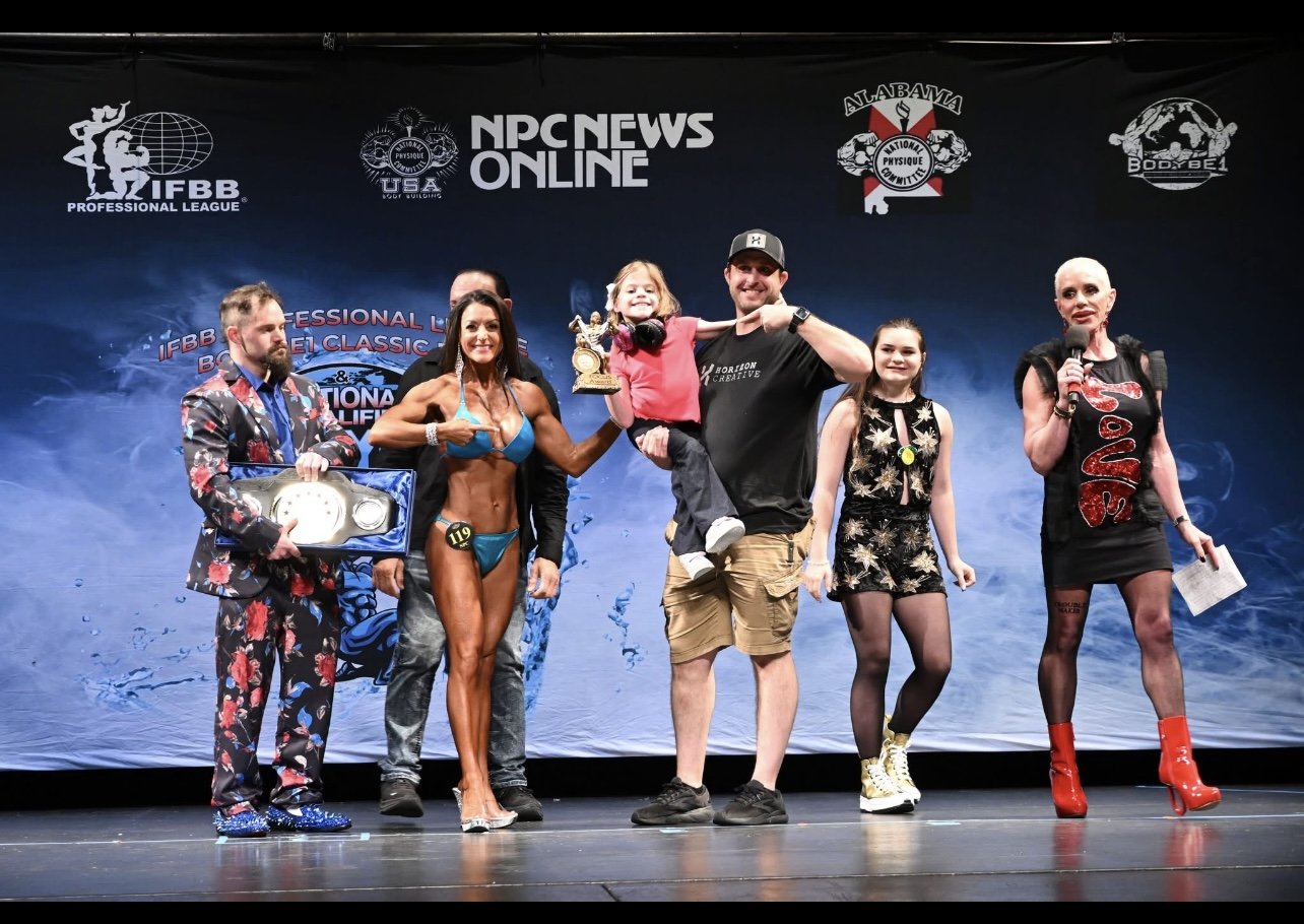 A group of seven people on a stage showcasing a bodybuilding competition, with some holding trophies and awards, and a backdrop displaying various fitness and bodybuilding logos.