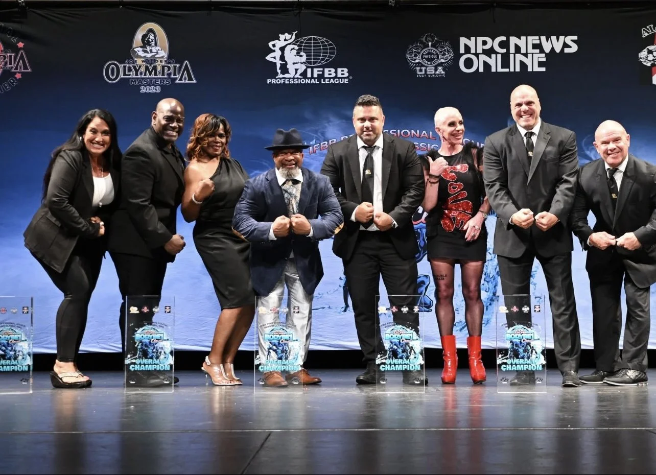 Group of eight people standing on stage during awards ceremony, with trophies labeled 'OverAll Champion' in front, at a bodybuilding or fitness event.
