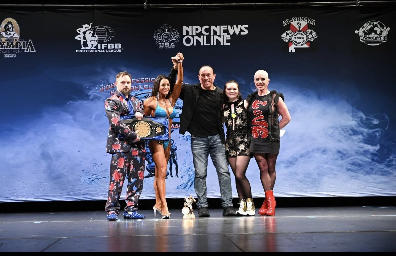 Group of five people standing on stage at a bodybuilding or fitness competition. One woman in a blue bikini holding a trophy, two women in stylish outfits, and two men, one in a floral suit and one in black, with one man raising a clenched fist. Back