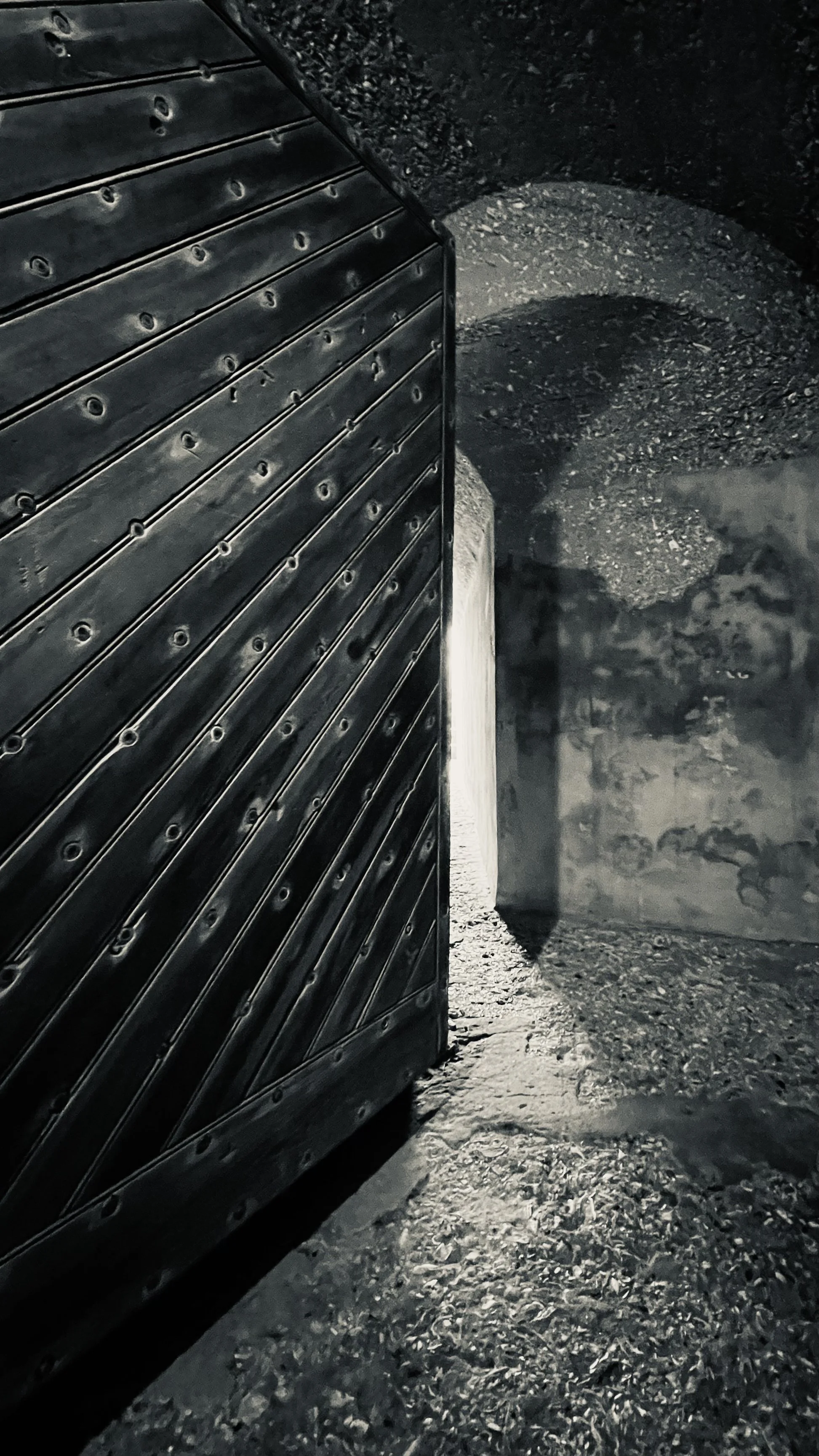 A black staircase with metal steps on the left side and a shadowed rough stone wall on the right side, with a concrete pillar in the middle and a dirt ground.