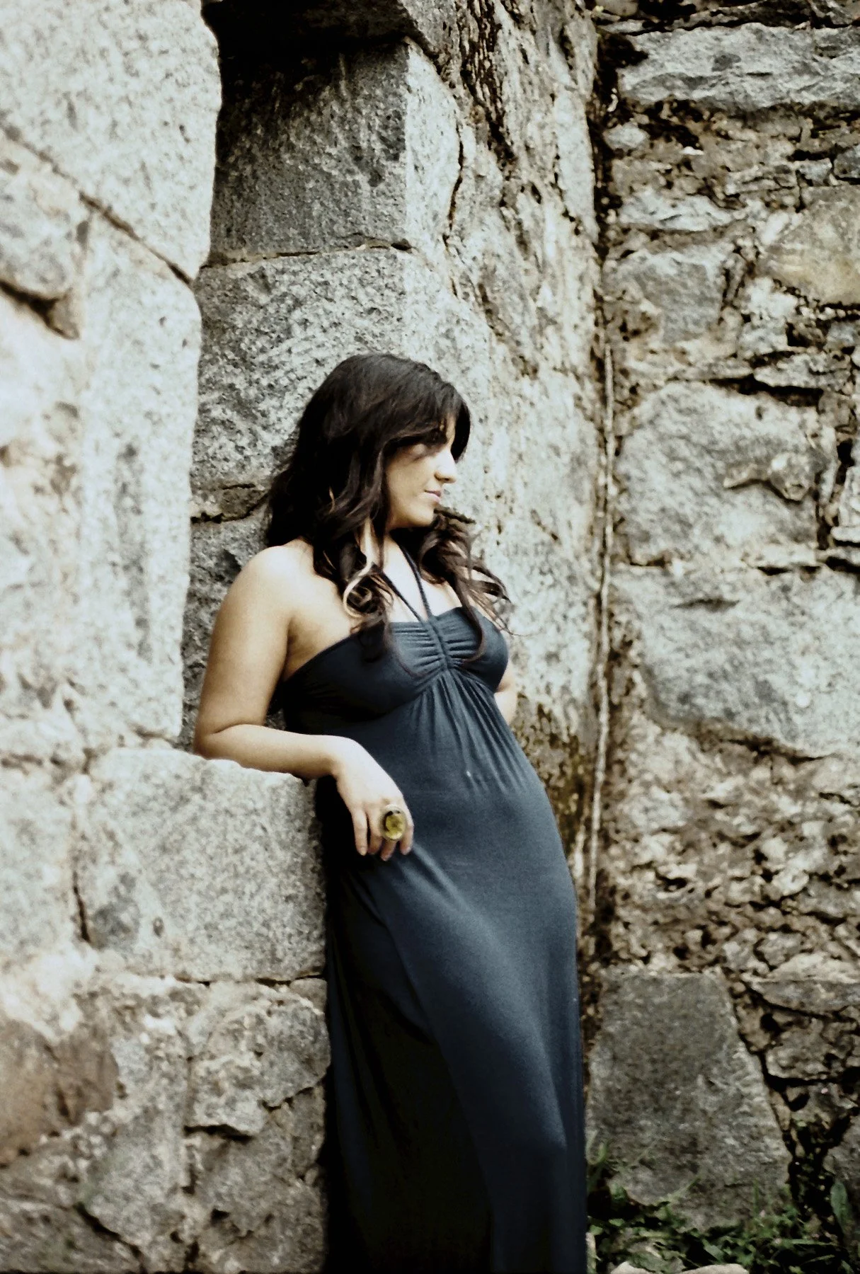 A woman with dark brown hair, wearing a black dress, stands against a stone wall, looking down with a serene expression.
