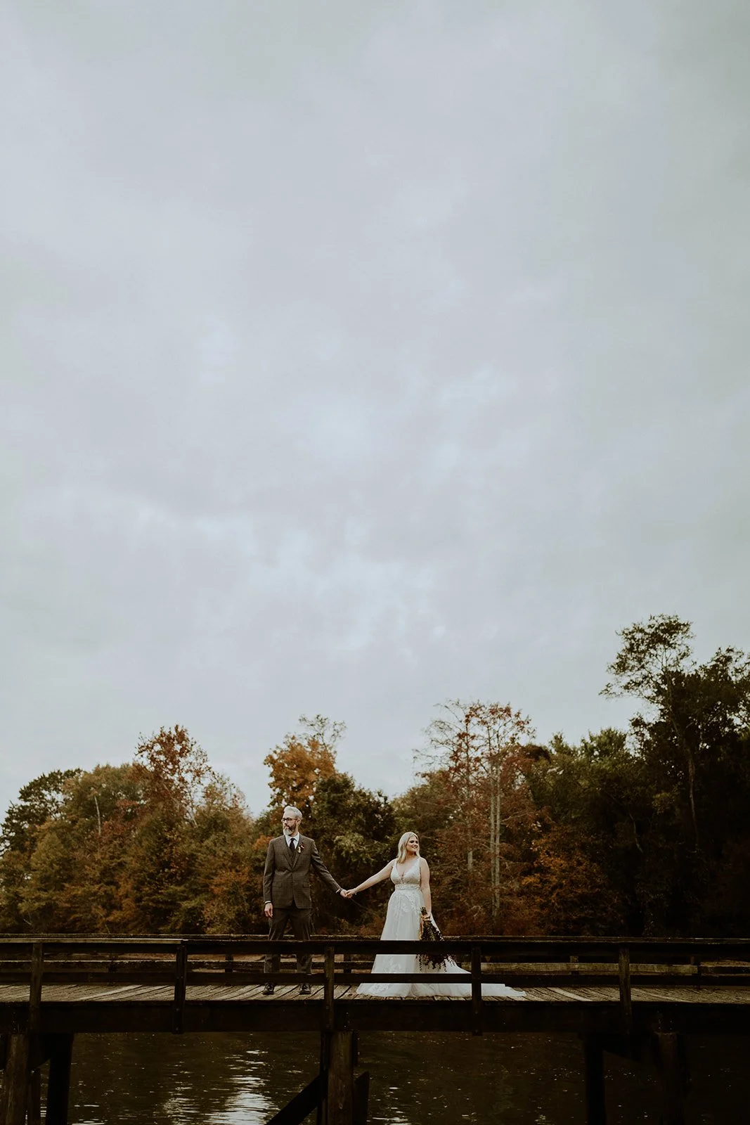 Bride and groom holding hands on a boardwalk with autumn trees in the background.