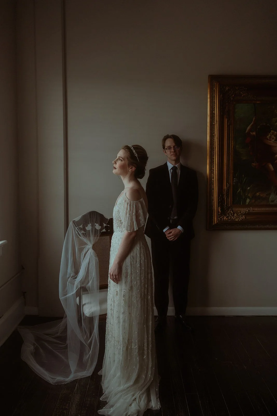 A bride in a white gown stands near a chair with a veil draped over it, looking upward. A man in a suit stands behind her, near a framed painting on the wall.