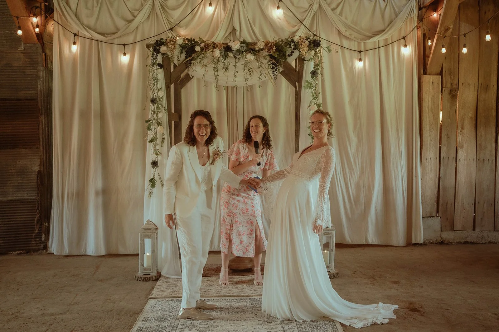 Three people in a wedding ceremony setup, two in wedding attire and one in a floral dress holding a microphone, standing under a decorated arch with curtains and string lights.