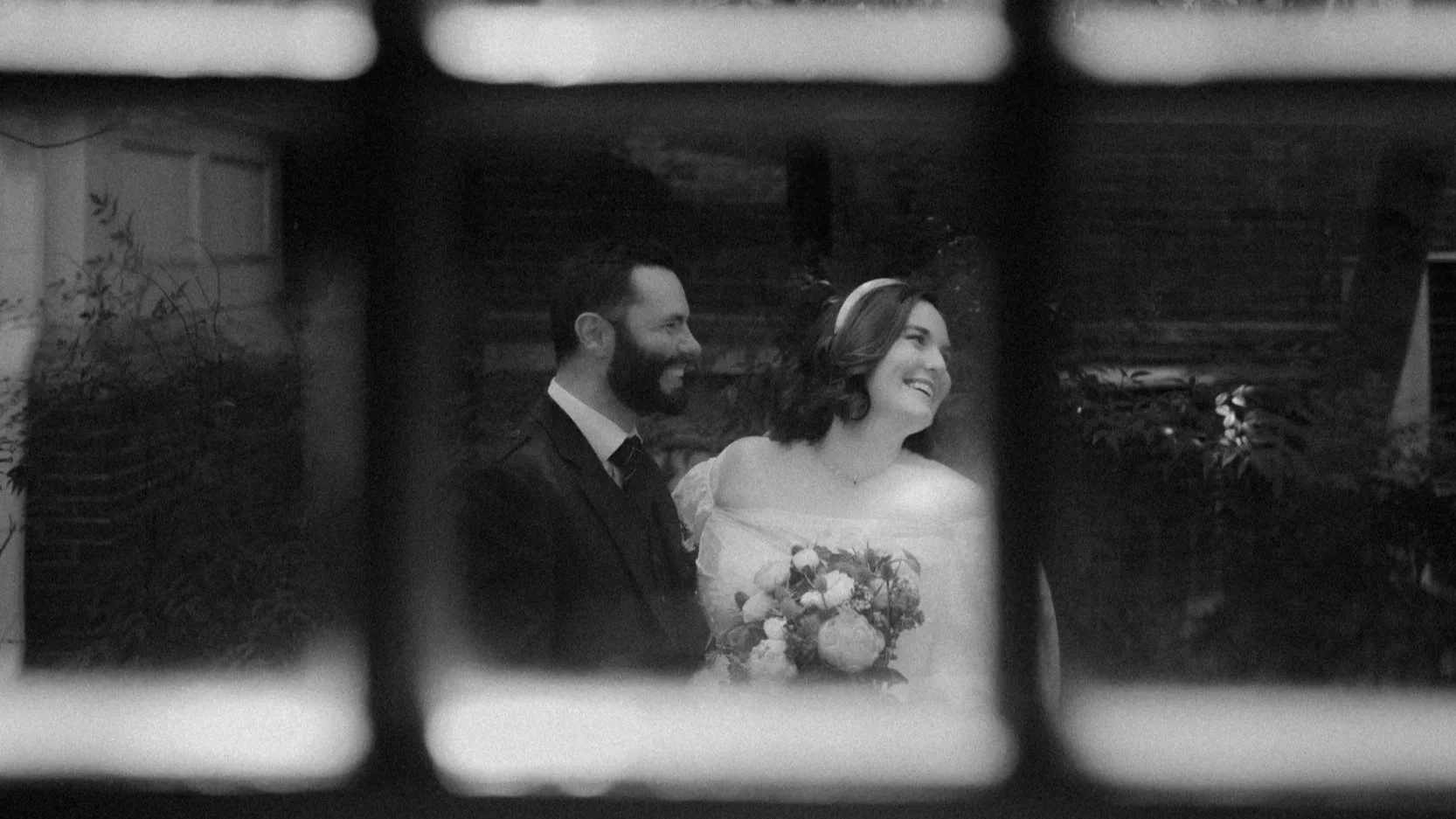 A black and white photo of a smiling bride and groom, seen through a window, on their wedding day.
