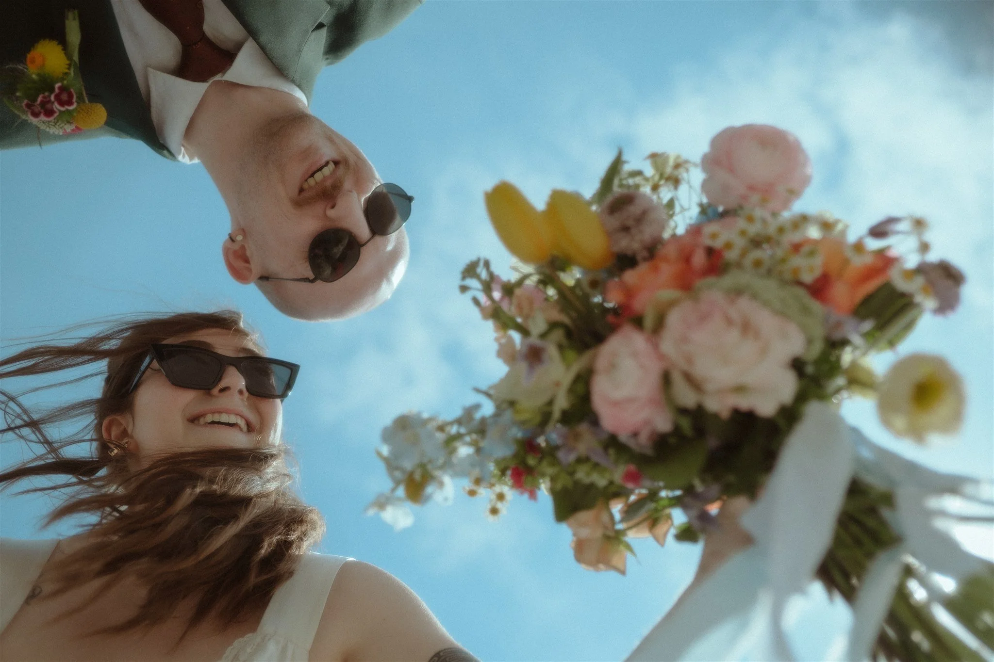 Two smiling people, a man and a woman, looking down at a colorful flower bouquet held by a person with long hair. They are wearing sunglasses and are outdoors on a sunny day.