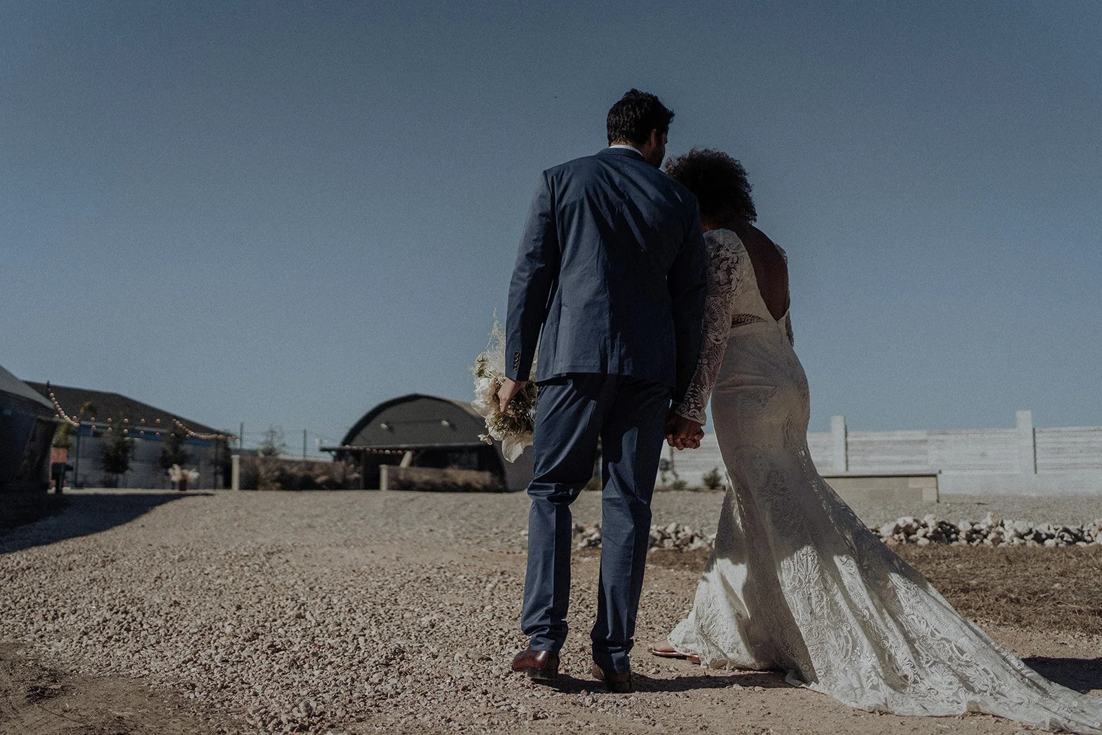 Bride and groom holding hands, walking on a gravel path against a clear sky.