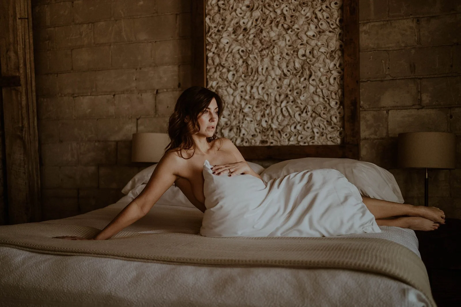 Woman wrapped in a blanket lying on a bed in a rustic room with textured wall art.