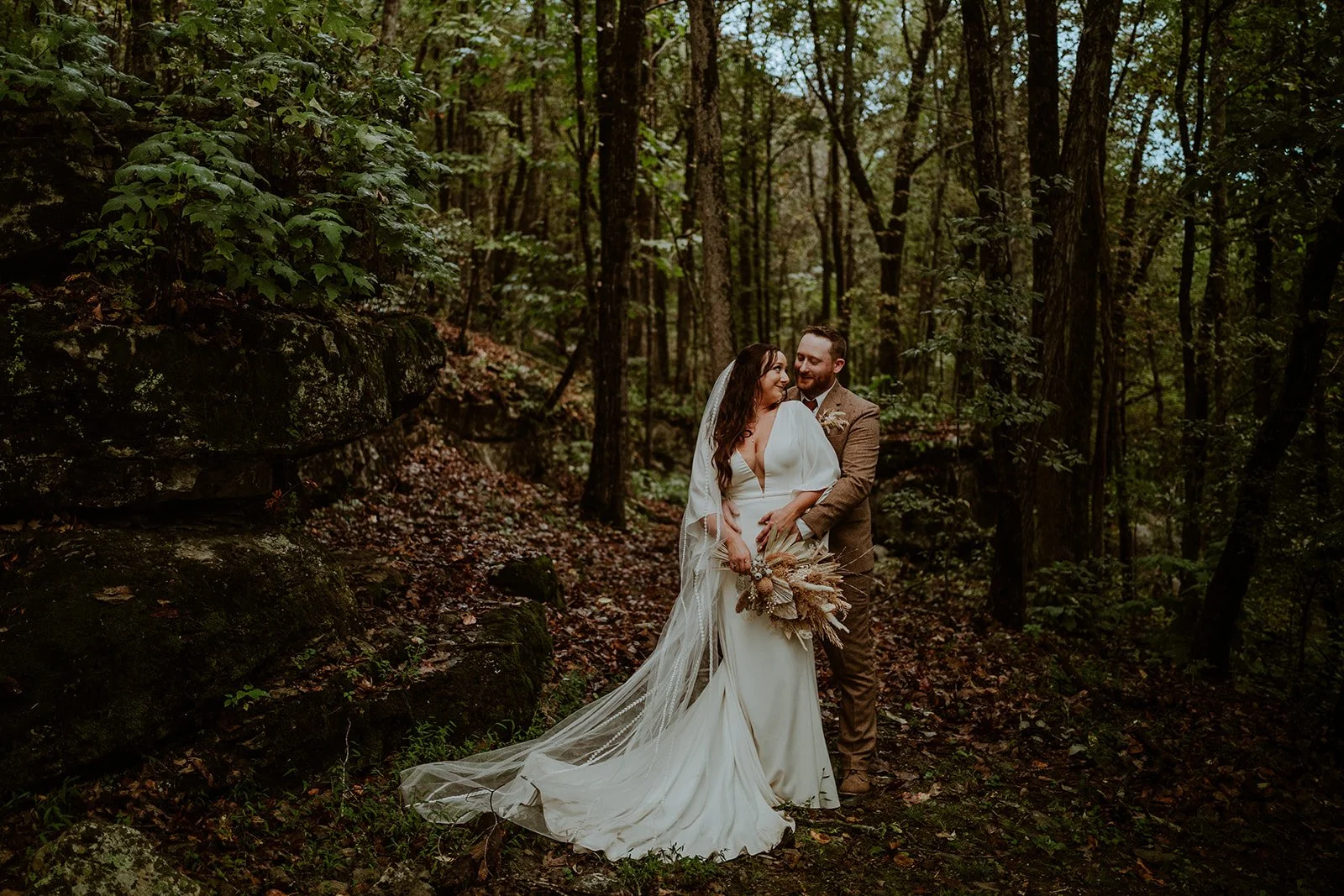 Bride and groom posing in a forest, with the bride in a white dress holding a bouquet, and the groom in a brown suit.