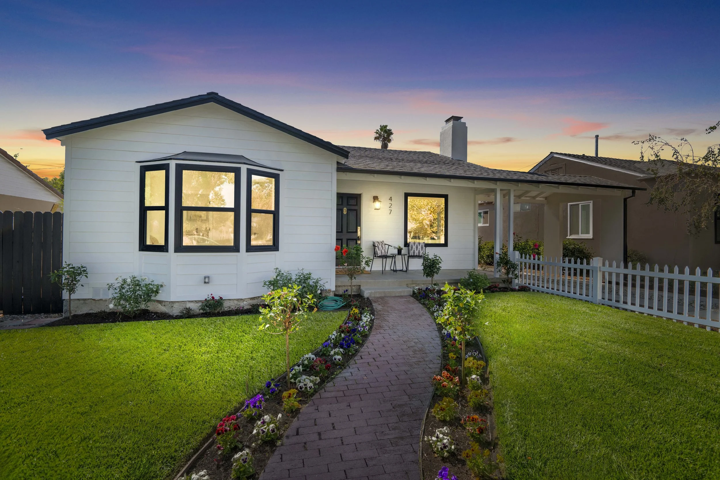 Sunset view of white suburban home with black window trim, flower-lined brick walkway, and cozy front porch — curb appeal captured by Intuition Real Estate Photography in LA.