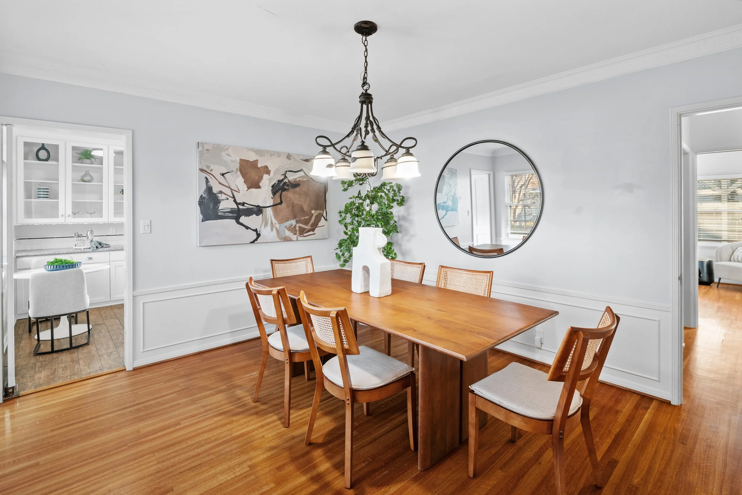 Dining room with wood table and six chairs, abstract artwork, chandelier, and wall-mounted round mirror — elegant staging captured by Intuition Real Estate Media in Los Angeles.