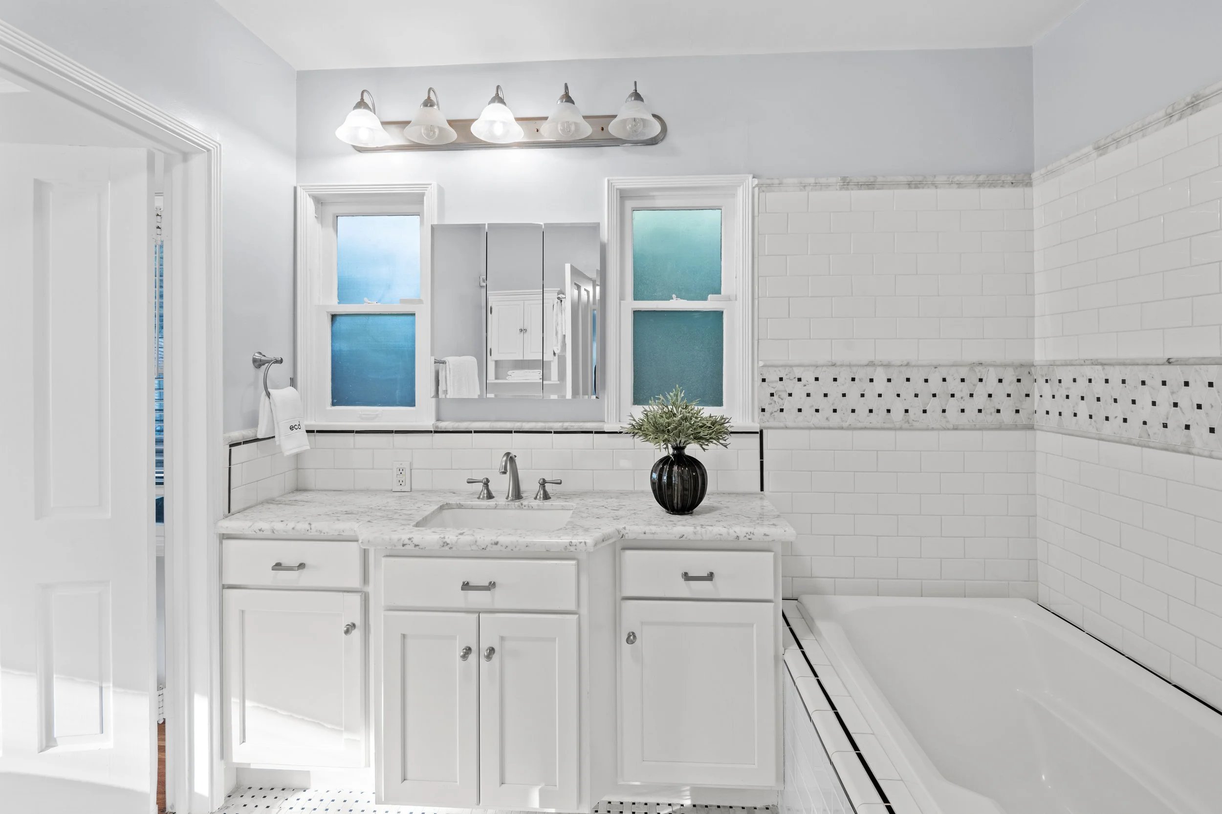 White bathroom with marble vanity, modern white cabinetry, frosted windows, and a soaking tub with white tile — real estate interior photo by Intuition Real Estate Media in Los Angeles.