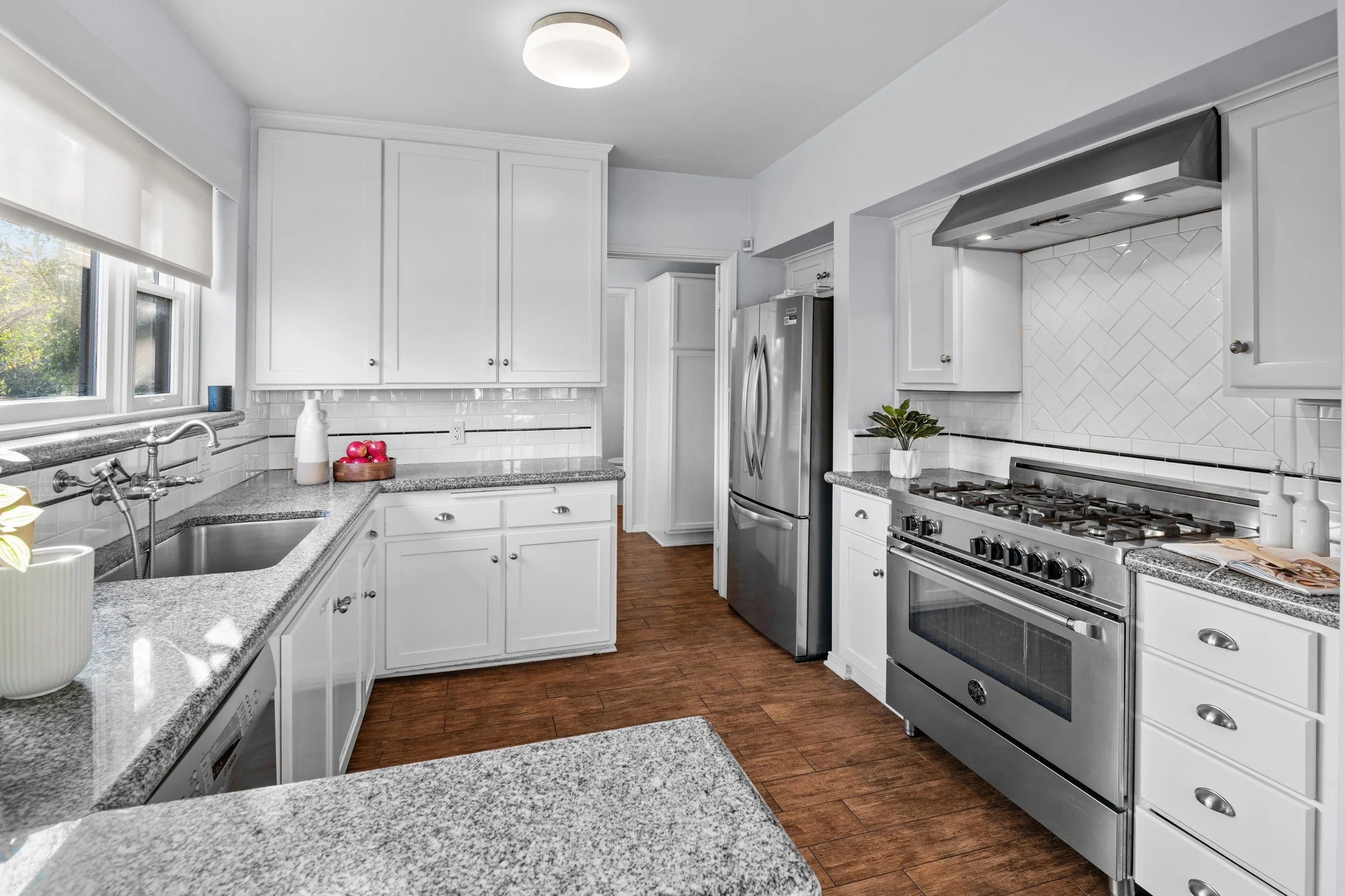 Bright white kitchen with granite countertops, stainless steel appliances, wood flooring, decorative plants, and a large window with a view of greenery — Los Angeles real estate photo by Intuition Real Estate Media.