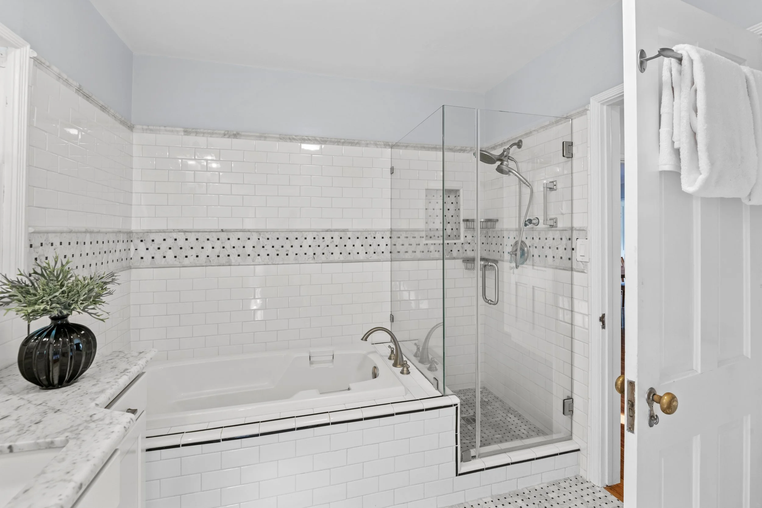 White bathroom with a glass shower, granite bathtub ledge, black and white mosaic tile border, and towel hooks — modern real estate listing photo by Intuition Real Estate Media in Los Angeles.