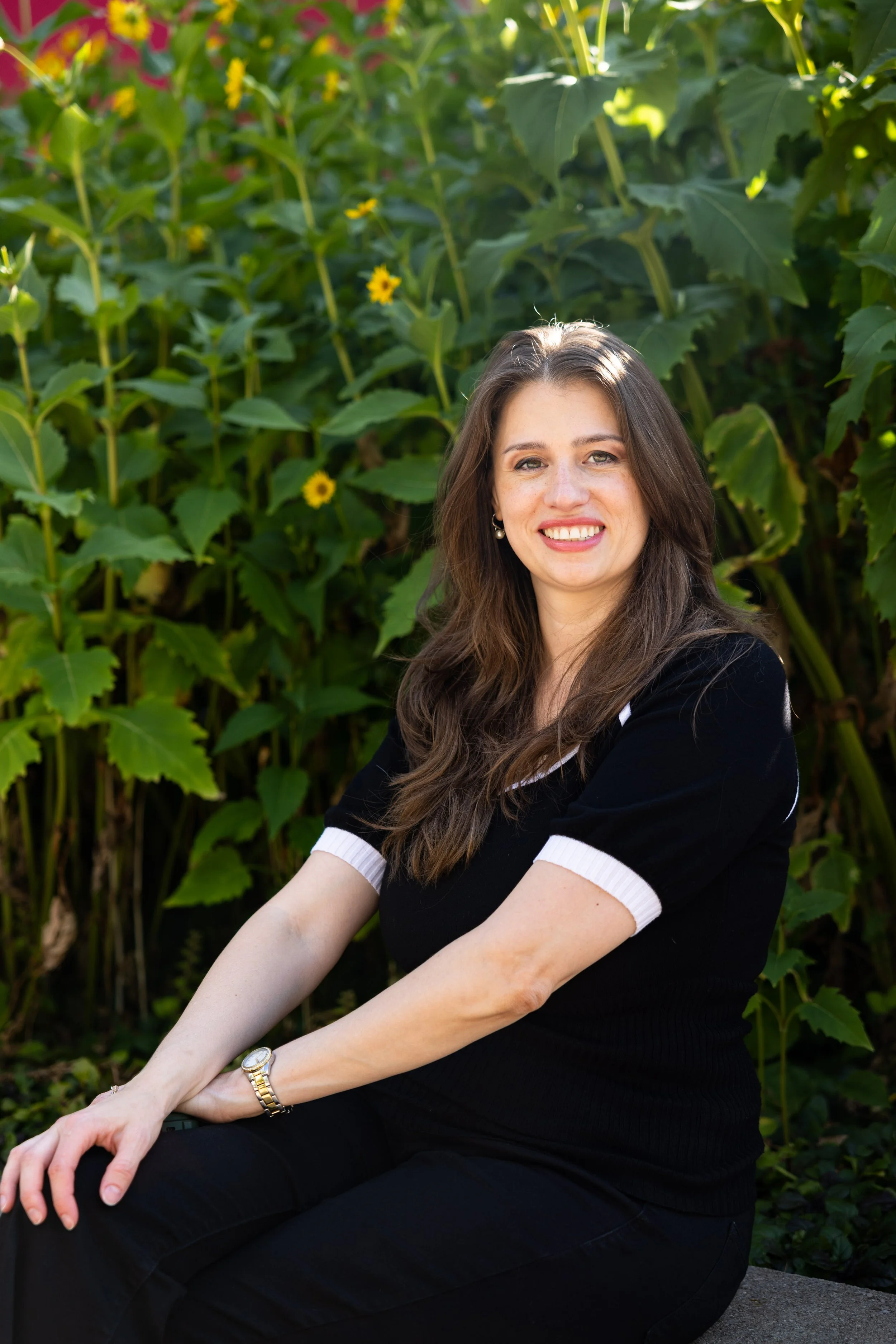 A woman with long brown hair and a black shirt with white cuffs, sitting outdoors against a background of green plants and yellow flowers, smiling at the camera.